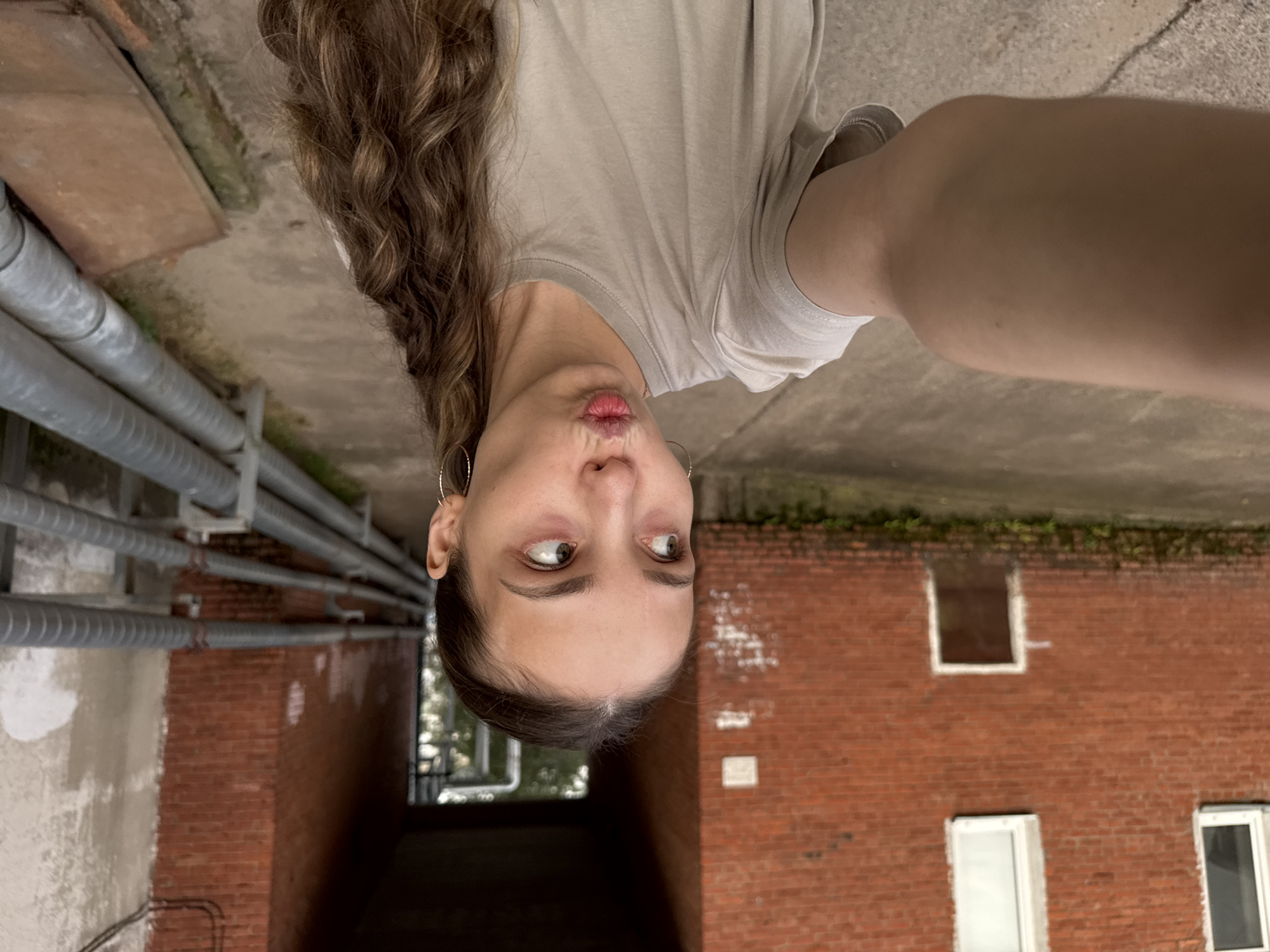 A young woman with long, curly brown hair and large hoop earrings takes a selfie in an outdoor urban setting. She is wearing a light tan t-shirt and puckering her lips. Behind her, a red brick building with white-trimmed windows and a small, dark door is visible. To her right, a walkway leads into a dark archway. Metal pipes run along the wall to the right of the walkway. The ground appears to be paved concrete with some mossy growth near the building.