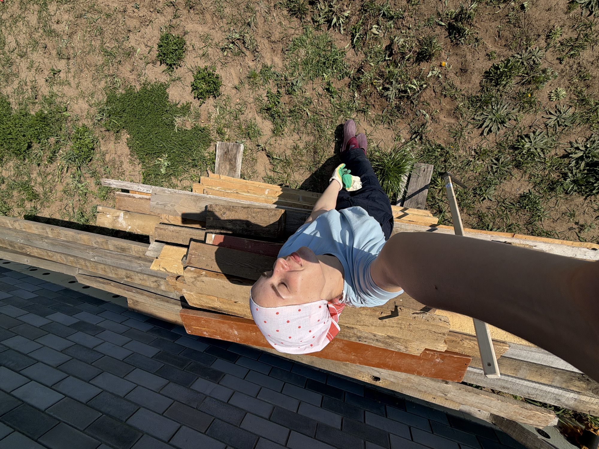 The image shows a woman lying on a pile of lumber. She is wearing a light blue shirt, dark pants, work gloves, and a white headscarf with red polka dots. The lumber is stacked on top of a paved surface. Beyond the lumber are patches of grass and bare earth. The photo appears to be taken in direct sunlight.
There is no information in the image that definitively confirms the location in Chaikovsky, Russia.