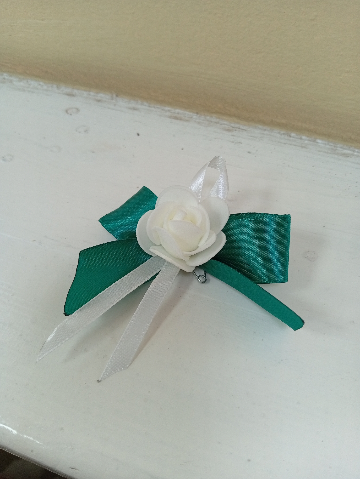 The image shows a white fabric rose attached to a green and white ribbon bow, secured with a pin. The item is placed on a white, textured surface, possibly a painted windowsill, with a light beige wall in the background. The surface appears slightly worn and has some small dark specks. The lighting suggests an indoor environment. The scene is located in Крыўлянскі сельскі Савет, Belarus.