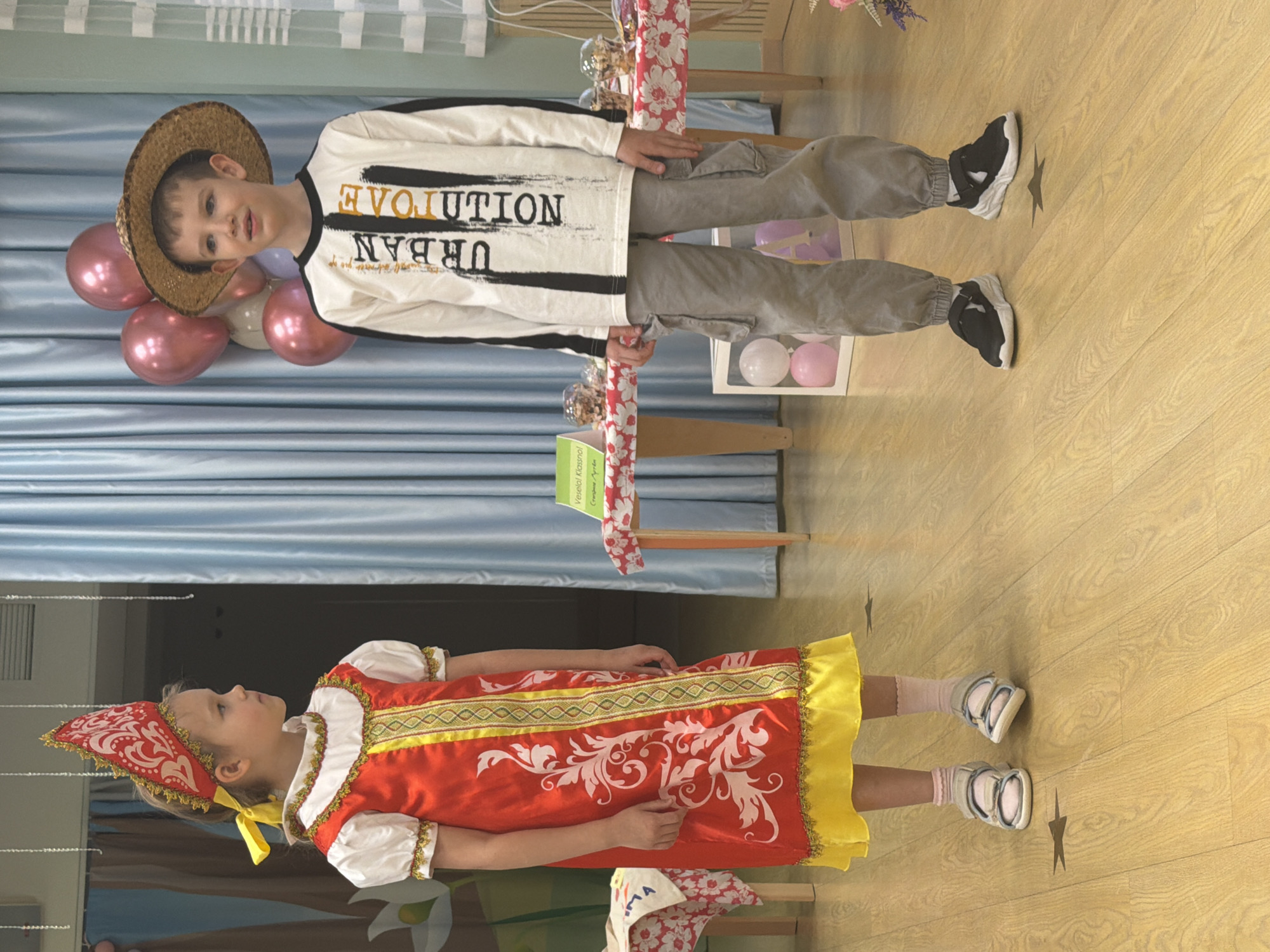 Two children are posing in a room with light-colored wooden flooring. A young girl wearing a traditional red and yellow dress with matching headwear is lying on the floor. A young boy wearing a hat, a long-sleeved shirt with the word "EVOLUTION" printed on it, and grey trousers is standing, holding a small table decorated with floral fabric. In the background, there is a light blue curtain and decorations. A framed sign is on the table, but the text is illegible. There are also balloons and a chair visible. There is no evidence to indicate the location is Saint Petersburg, Russia.