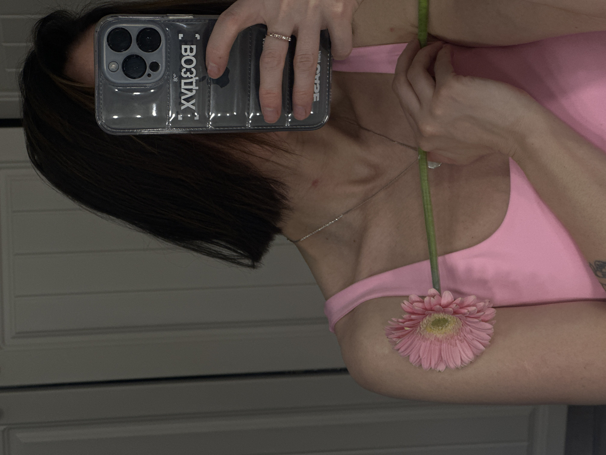 A woman is taking a selfie with her phone, which has a transparent case with the word "ВОЗДУХ" written vertically on it. The phone is held in her left hand, and she's wearing a ring. She's holding a pink gerbera daisy in her right hand, and the flower's head rests on her right shoulder, which is covered by a pink top. She is wearing a thin necklace. Her dark, short hair is visible behind the phone. The background is a white, paneled wall. There's no evidence indicating the location to be Votkinsk, Russia.