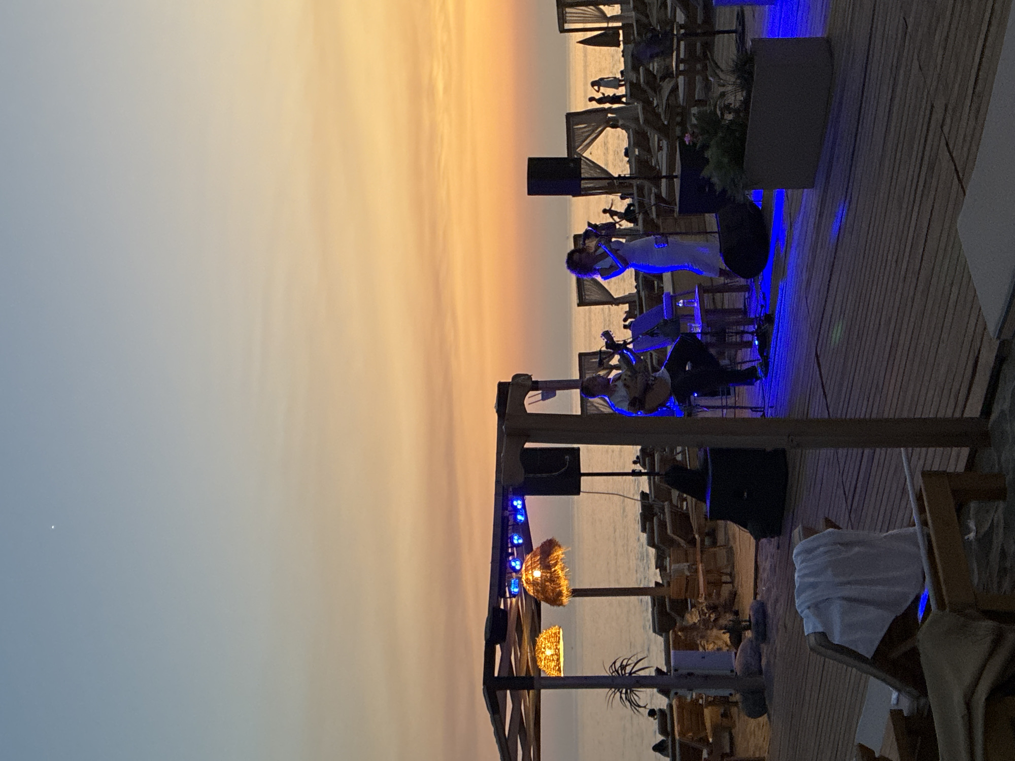 The image captures a beachside performance at dusk in Alanya, Turkey. A male musician plays an acoustic guitar on a wooden stage, accompanied by a female singer in a white dress who is holding a microphone. Blue stage lights illuminate the performers and the surrounding wooden deck. In the background, the Mediterranean Sea is visible, with lounge chairs and cabanas set up on the beach. The sky displays a gradient of soft orange and blue hues, typical of a sunset. Several people are scattered across the beach, enjoying the ambiance. The scene suggests a relaxed evening at a beach club or restaurant.