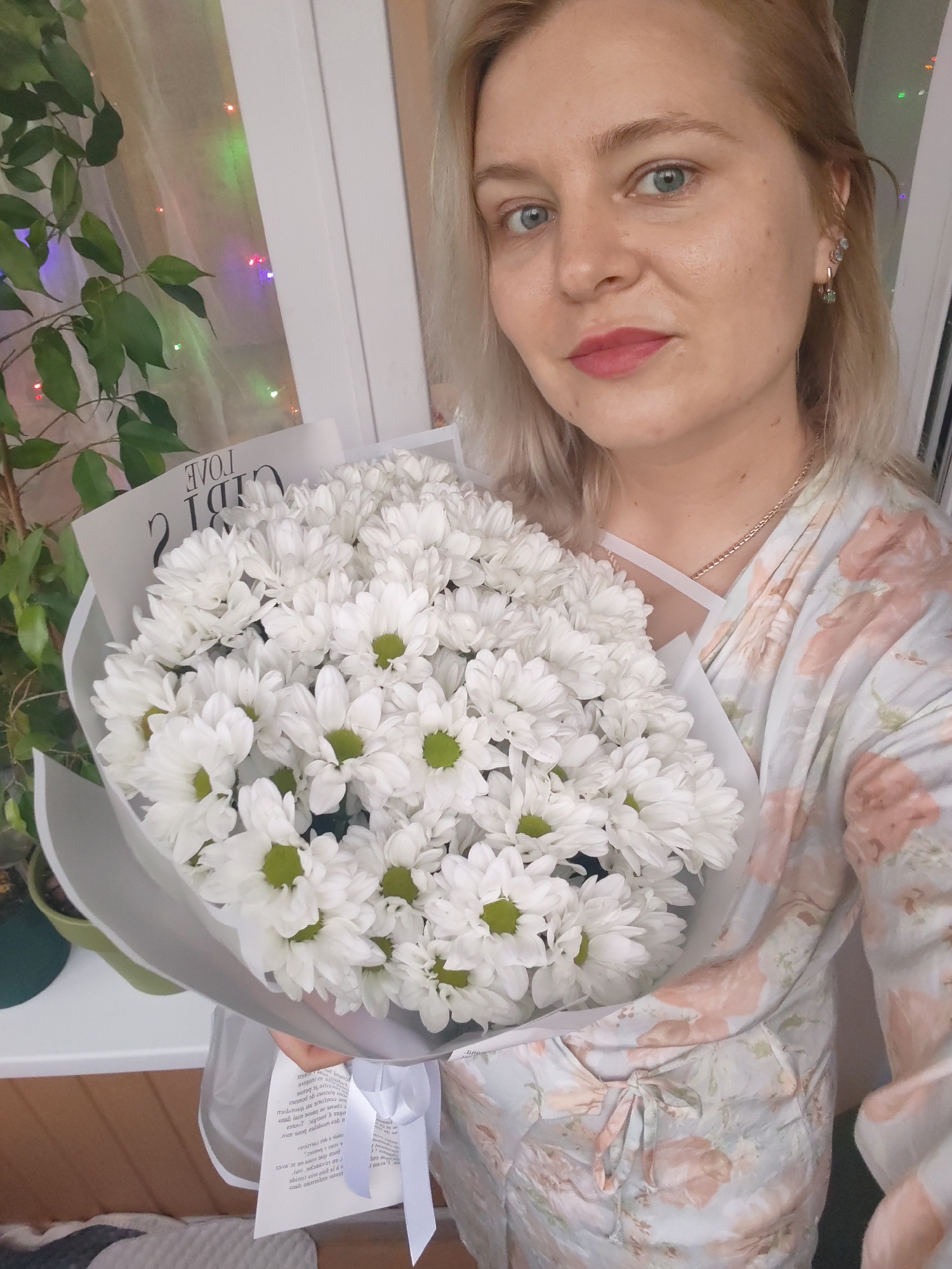 This media file shows a close-up, selfie-style shot of a young woman holding a large bouquet of white chrysanthemums. The woman has blonde hair, light skin, and is wearing pink lipstick and light eye makeup. She has a light blue, floral-patterned robe or dress on, and is wearing a delicate gold necklace. She is also wearing earrings with small blue stones.
The bouquet is the dominant object in the foreground, with the white petals and green centers of the flowers clearly visible. The flowers are wrapped in translucent white paper and tied with a white ribbon. A tag with text is attached to the bouquet.
In the background, to the left of the woman, is a large green plant with broad leaves. Behind the plant and the woman, there is a window with colorful lights (red, green, blue, yellow) visible, suggesting it might be evening or a festive occasion. The overall lighting suggests indoor illumination.
Visible text on the tag includes Cyrillic letters, with words like "CIỜI C" and "ΓΟΛΕ" being partially legible. The text appears to be a mix of Cyrillic and possibly Latin characters, suggesting a decorative or brand element associated with the bouquet.
The woman's expression is neutral to slightly smiling, and she appears to be posing for the picture, holding the bouquet up to display it. The setting appears to be a home environment, given the presence of the plant, window, and possibly indoor decorations. The date context is not explicitly provided within the image itself, but the general ambiance is casual and celebratory.
