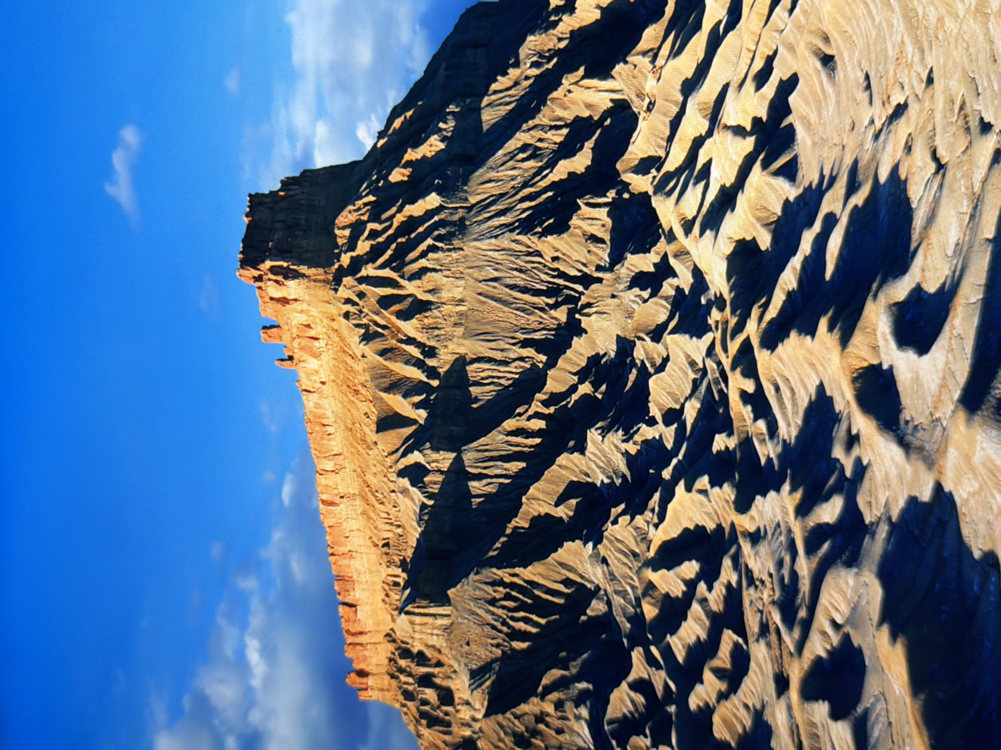 The image depicts a dramatic desert landscape with a prominent mesa or butte bathed in warm, golden light under a vibrant blue sky. The geological formations are characterized by steep, eroded slopes with deep shadows, creating a stark contrast between illuminated and shaded areas. The top of the mesa is flat and appears to be layered rock, with some smaller rock outcrops. The foreground shows a rugged, undulating terrain with similar erosional patterns, suggesting a dry, arid environment. The sky is mostly clear with a few scattered white clouds. There are no people, animals, or man-made structures visible in the image. The location is a natural desert environment, not a city.