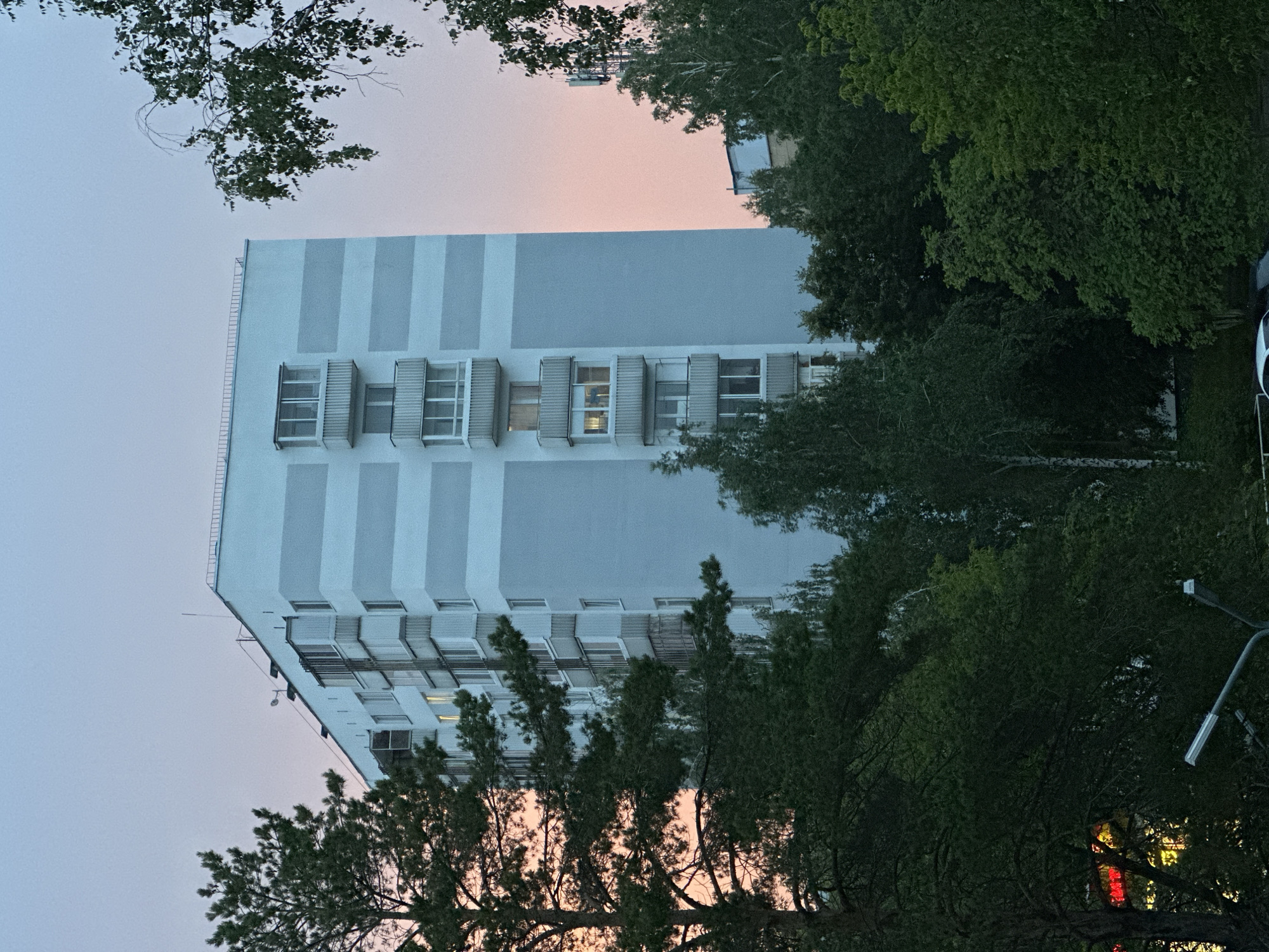 The image shows a tall, modern apartment building with a light blue and white facade. The building has multiple floors with rows of windows, some of which appear to be balcony doors. Vertical white stripes alternate with blue panels on the building's exterior, creating a striped pattern. Surrounding the building are dense green trees, obscuring parts of the lower floors and the ground. The sky above is a gradient of soft pink and pale blue, suggesting either sunrise or sunset. A few trees are in the foreground, their branches and leaves framing the view of the building and sky. There are no people visible in the image. The scene appears to be residential, possibly in a suburban or urban setting. The lighting and the colors in the sky indicate it is either early morning or late evening. No text is visible in the image.