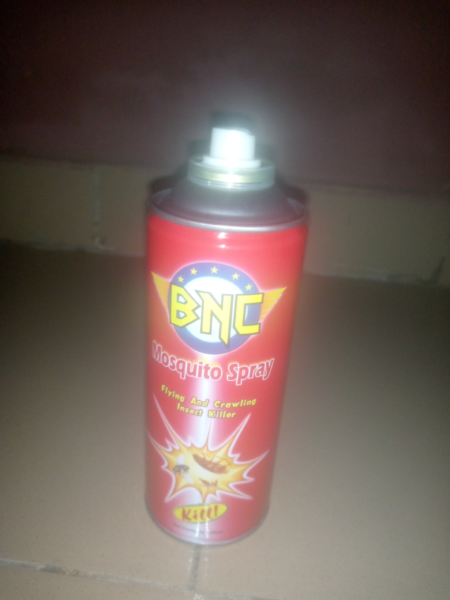 A single red cylindrical aerosol can of "BNC Mosquito Spray" stands upright on a light-colored tiled floor in a dimly lit indoor environment, located in the city of Unknown, Unknown. The can features a yellow "BNC" logo with a lightning bolt and four yellow stars on a blue and white circle. Below the logo, the text "Mosquito Spray" is visible, followed by "Flying And Crawling Insect Killer." Near the bottom, a graphic depicts a mosquito and a cockroach within a yellow burst, with the word "Kill!" printed beneath it. The can has a silver-colored top with a white plastic spray nozzle. The background consists of a dark, undifferentiated surface, likely a wall, appearing out of focus.