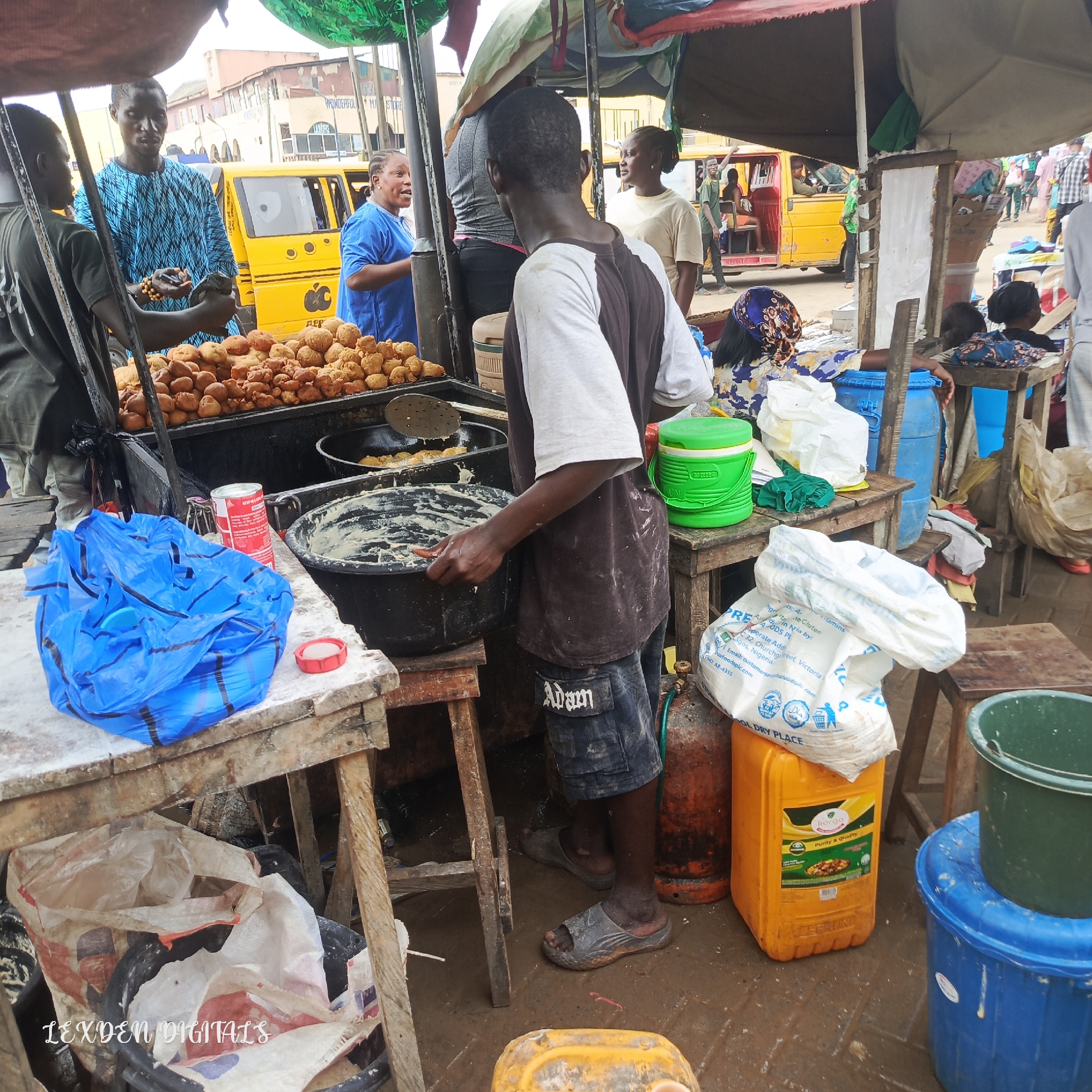 This media file captures a bustling street scene in Egan, Nigeria, likely during the day. The primary focus is on a food vendor preparing and selling what appear to be fried snacks. In the foreground, a man is actively engaged in preparing the food, stirring a large pot of batter with a spoon. Piles of golden-brown fried snacks are prominently displayed on a table in front of him, suggesting a busy market or roadside stall.
Several other individuals are present in the scene. A man in a blue patterned garment stands near the food stall, engaged in conversation or observation. A woman in a blue shirt is also interacting nearby, her expression lively. In the background, a yellow commercial vehicle, common in Nigeria, is visible, suggesting a public thoroughfare or transport hub. Other people are scattered throughout the scene, some walking or standing, contributing to the sense of a busy marketplace.
Various objects are scattered around the food stall, including plastic bags, buckets of different colors (blue, green, orange), and what appears to be a gas cylinder. The ground is wet and somewhat muddy, hinting at recent rain or general market conditions. The overall atmosphere is one of commerce and daily life, with a focus on food preparation and sale. The visible text includes "LEXDEN DIGITALS," "WONDERFUL MA STORE," and text on packaging that mentions "Purity & Quality," and nutritional information, indicating the location of the stall and potentially the origin of some products. The time of day is suggested to be daytime by the lighting.