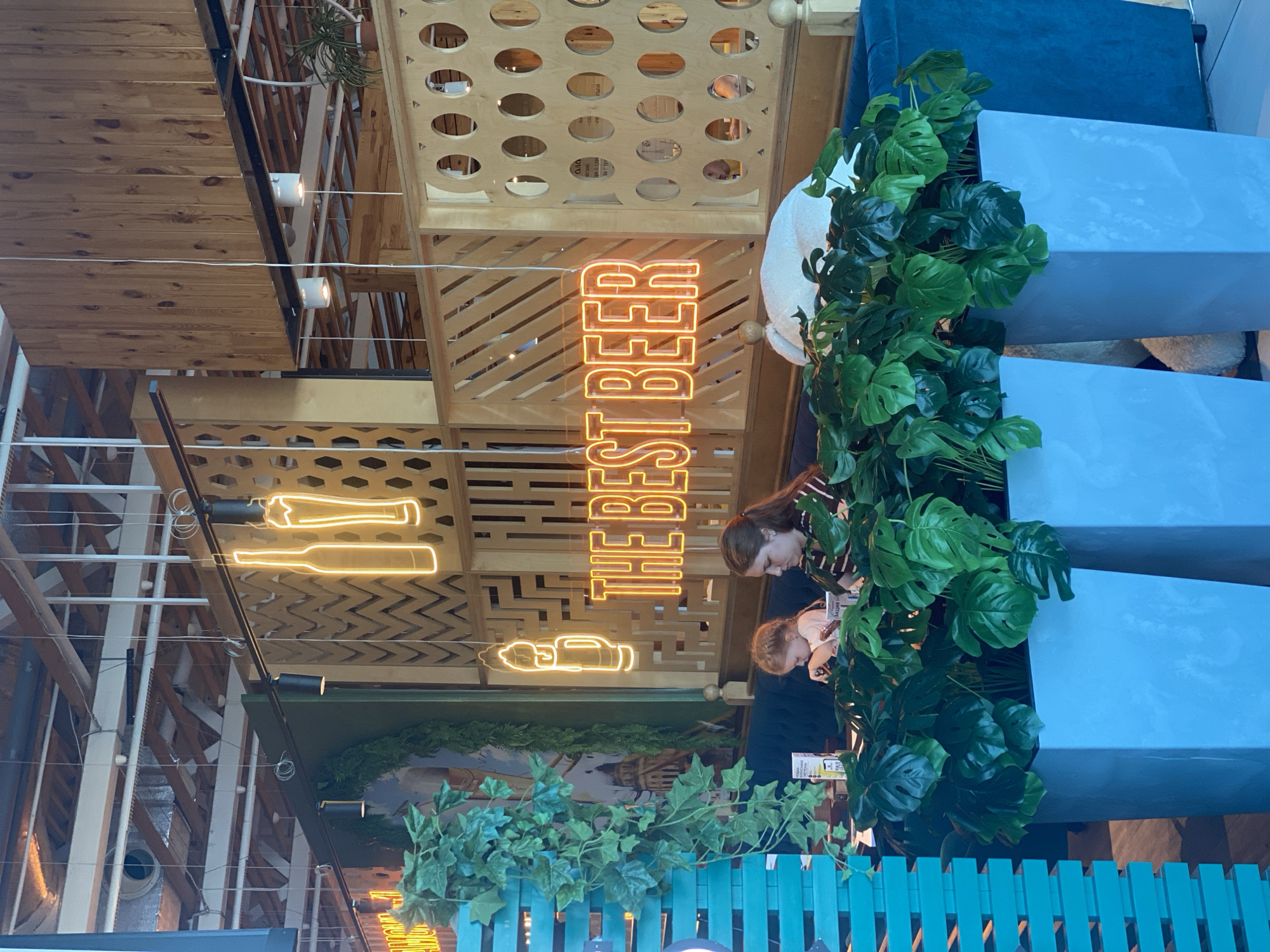 The image shows the interior of a restaurant, likely "The Best Beer," located in Ob, Russia. A neon sign with the words "THE BEST BEER" glows in orange. Two people, a woman and a young girl, are seated at a table in the foreground, appearing to be eating. Lush green plants in large blue planters are in front of them, partially obscuring the view of the lower half of the table. The restaurant's decor features wooden elements, hexagonal patterns, and hanging lights. The overall ambiance is casual and modern, suggesting a relaxed dining experience.