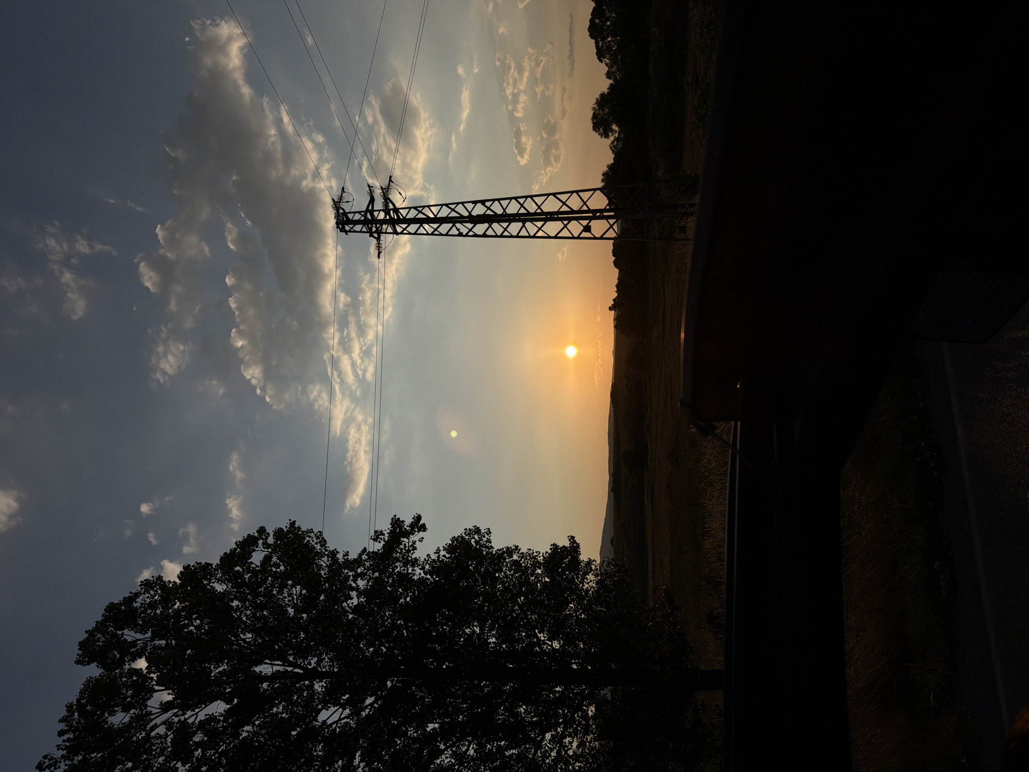 A silhouette of a large tree stands on the left side of the image against a sunset sky. In the mid-ground, a power pylon is visible, with power lines stretching across the frame. The sun is low on the horizon, casting a warm glow across the sky and landscape. The foreground is largely obscured by a dark silhouette, possibly a car interior or a roadside barrier, with the dark outline of a road visible at the bottom. The scene appears to be in a rural or semi-rural area, with fields and distant hills in the background. The location is identified as Bogdanovdol, Bulgaria.