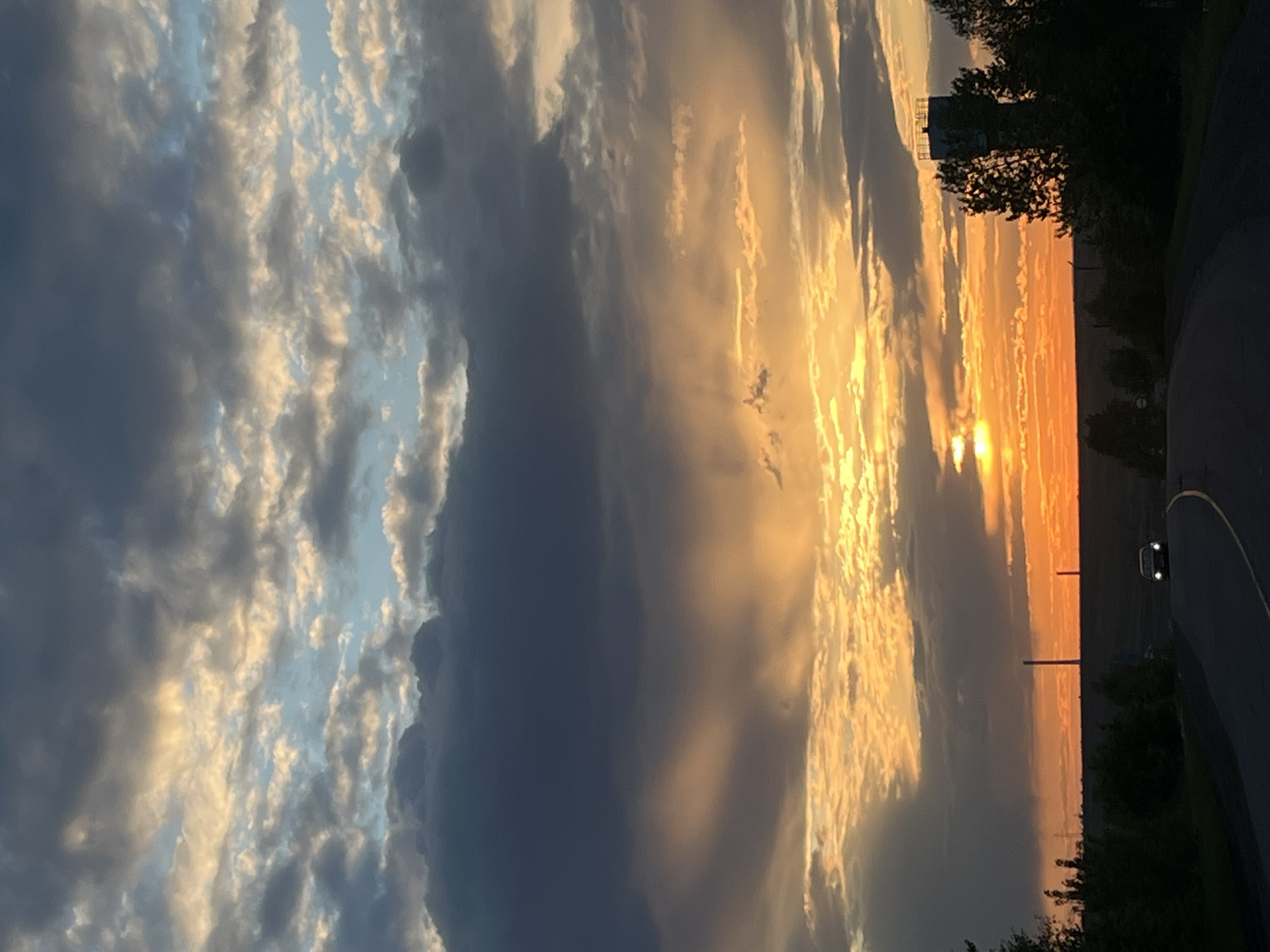 The image shows a sunset in Зыково, Russia. The sky is filled with clouds, with a mix of dark gray and light blue-white patches. The sun is setting on the horizon, casting a golden orange glow across the landscape. There is a road with a car on the right side of the image, with trees and vegetation bordering the road. In the distance, thin vertical structures are visible on the horizon.