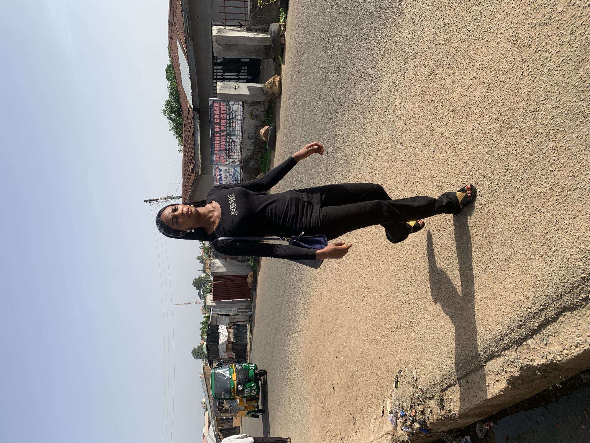 The image shows a woman wearing a black outfit and sandals, standing on a paved road in Jos, Nigeria. She has a small blue purse slung over her shoulder. In the background, there are buildings, one of which has a sign that reads "THE THRONE OF GRACE INT'L GOSPEL MINISTRY," indicating a religious location. A yellow and green auto rickshaw is visible on the side of the road, typical of transportation in this area. The road surface is worn and the surrounding area shows signs of urban life with various structures and objects scattered around.