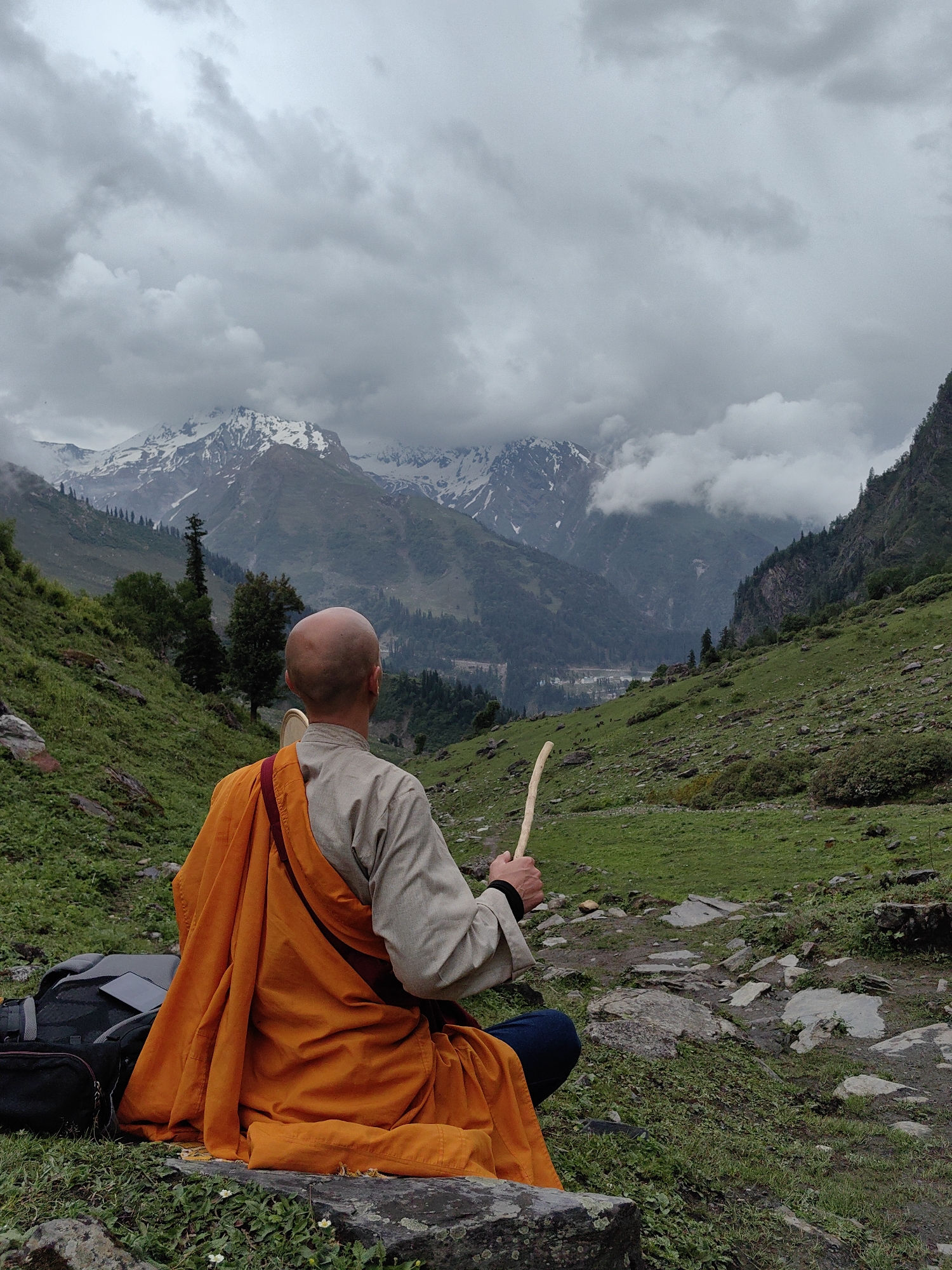 A man wearing an orange robe and a gray shirt is sitting in a meditative pose on a rocky path in a mountainous region. He has a shaved head and is holding a wooden stick in his right hand. A black backpack is next to him. The background features lush green hills, rocky terrain, and snow-capped mountains under a cloudy sky. The image captures a scene from the city of Dhundi, India, known for its natural beauty and trekking routes.