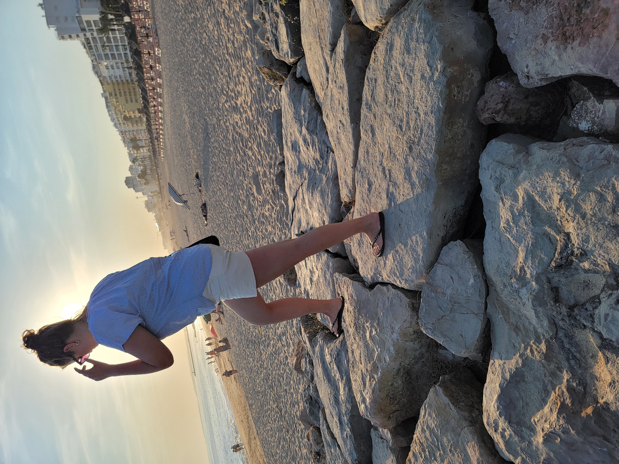The image depicts a young woman standing on a rocky embankment next to a sandy beach. She is wearing a light blue striped short-sleeved shirt, cream-colored shorts, and black flip-flops. Her hair is pulled up in a bun, and she is holding pink-rimmed sunglasses. She is looking downwards, seemingly adjusting them.
The background shows a beach with people walking, and buildings line the edge of the beach in the distance. The ocean is visible to the left. The scene appears to be set during sunset, as indicated by the warm, golden light illuminating the rocks and the soft glow in the sky. The weather appears to be clear and pleasant.
The rocks are large and uneven, with patches of dry grass growing between them. The sand has visible ripples from the receding tide or wind. The overall impression is a relaxed, end-of-day scene at a coastal location. The location context is Quarteira, Portugal. There is no visible text in the image.