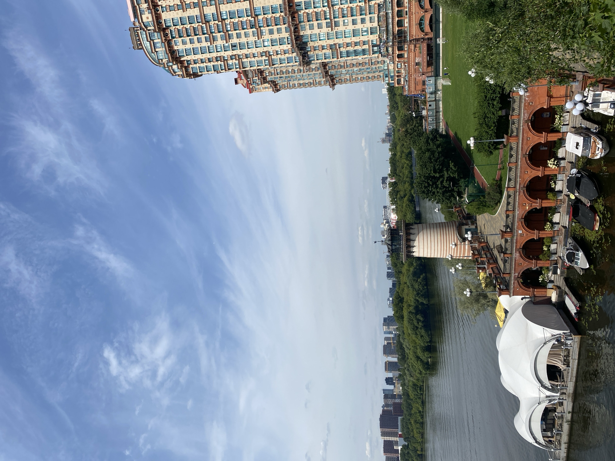 This is a vertical, medium-shot, eye-level photograph of a waterfront scene in an urban environment under a bright, partly cloudy sky. In the foreground, a large white tent structure with arched openings sits on a dock on the left side of the frame, appearing to be a restaurant or event space. Several boats are moored to the dock. To the right of the tent, a red brick building with arched windows and balconies stretches along the waterfront. Above this building, a lighthouse-like structure with horizontal stripes is visible. A body of water, likely a river, occupies the bottom left quadrant of the image, with gentle ripples on its surface.
In the mid-ground, a green lawn with a soccer field is visible, bordered by trees. The opposite bank of the river is lined with a dense forest and a skyline of numerous residential and commercial buildings. The sky is a light blue with scattered white cirrus clouds, some wispy and others more defined. A large, modern apartment building with a distinctive curved facade and many windows dominates the right side of the frame, extending from the mid-ground to the top. The overall impression is of a well-developed urban waterfront with a mix of modern and older architectural styles.