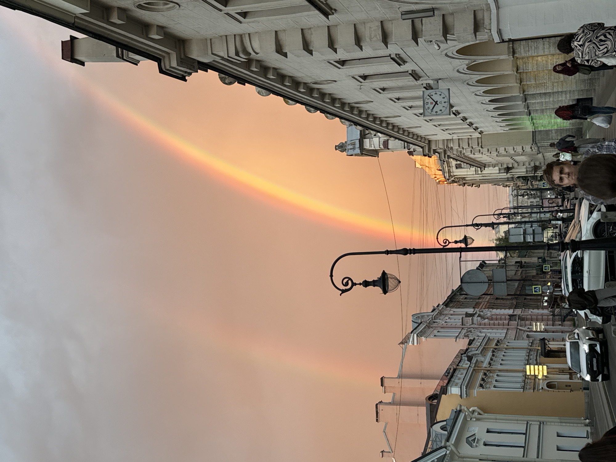 This image depicts a street scene in Saint Petersburg, Russia, characterized by its architectural style and the presence of a prominent rainbow. The sky is a soft, warm gradient of orange and pink, suggesting either sunrise or sunset. A faint double rainbow arcs across the sky, its colors muted by the ambient light.
The street is lined with historic buildings featuring ornate facades, arched windows, and decorative elements. Lampposts with classic designs are visible along the street, adding to the historical ambiance. Several cars, including white sedans, are parked or driving on the road. A few people are walking along the sidewalk, some with shopping bags, suggesting a casual outing. One person is looking directly at the camera.
A large clock is mounted on one of the buildings, indicating the time. The overall atmosphere is serene and perhaps a bit melancholic, possibly due to the soft lighting and the fading light of day. The presence of the rainbow adds a touch of wonder and beauty to the urban landscape. The image captures a fleeting moment of natural beauty against the backdrop of a historical city.