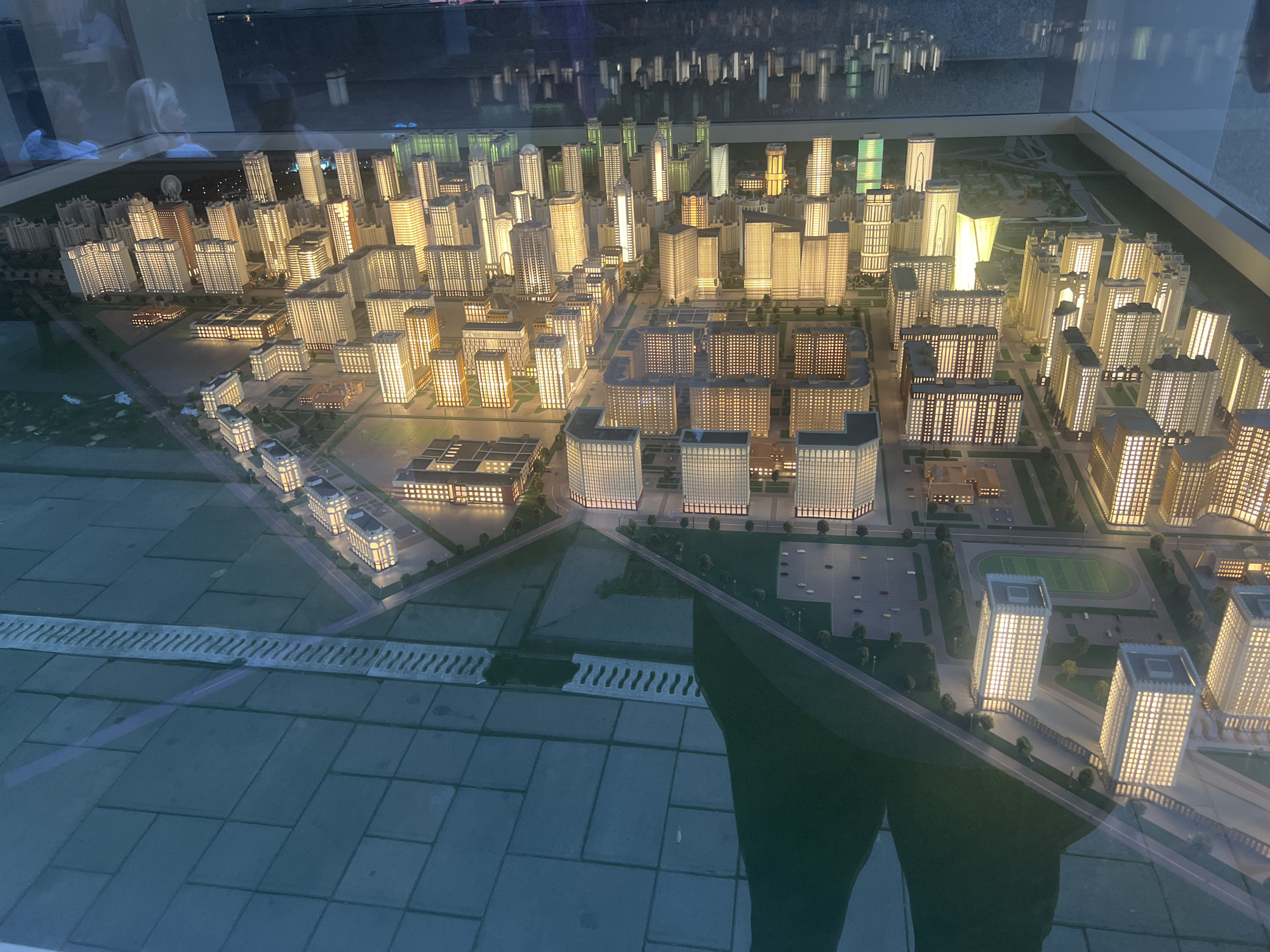 The image displays a detailed architectural model of a city district, illuminated at dusk or night. Numerous multi-story buildings, representing residential and commercial structures, are lit from within, creating a vibrant urban landscape. The model includes roads, green spaces, and what appears to be a sports field with a running track. In the foreground, a textured surface with a drainage grate is visible, suggesting a display base. The overall impression is of a modern, well-developed urban area. While the prompt mentions Grozny, Russia, this cannot be confirmed from the image alone.