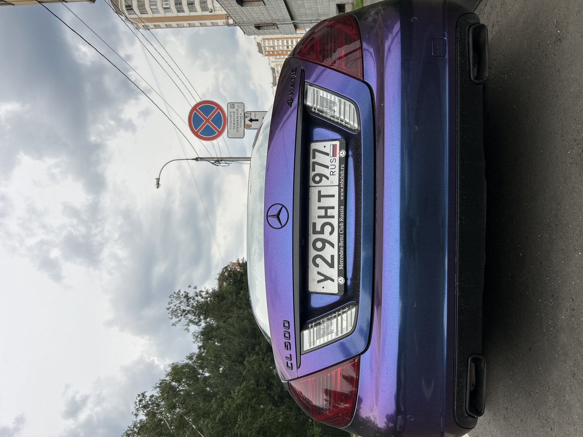 A Mercedes-Benz CL 500 with a unique purple and blue iridescent paint job is parked on an asphalt road. The car's rear is facing the camera, displaying its license plate which reads "y 295HT 977 RUS". Below the license plate, the text "Mercedes-Benz Club Russia" and "www.mbclub.ru" is visible. The car is situated in front of a building with a gray facade and an apartment building with many windows in the background. A street sign showing a red circle with a blue cross indicates no stopping, and another sign below it has Russian text and an arrow pointing upwards. Trees line the left side of the road, and power lines are visible overhead. The sky is overcast with gray clouds.