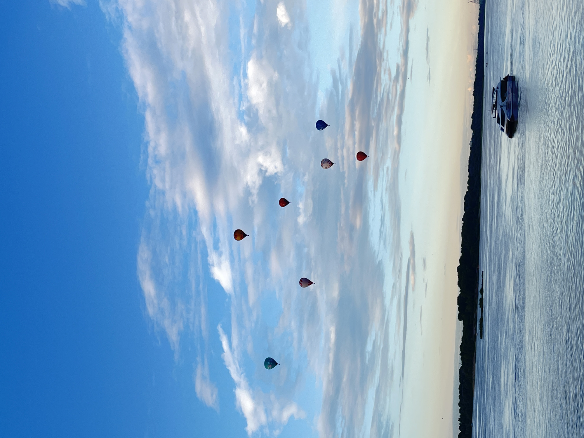 This media file captures a serene scene of hot air balloons floating in a vast blue sky dotted with wispy clouds. There are seven hot air balloons visible, each a distinct color, adding pops of vibrancy against the pale sky. In the foreground, a body of water, likely a river, stretches across the frame, its surface rippling gently. The far bank of the river is lined with a dark, dense forest. A modern, dark-colored boat is cruising on the water, its silhouette contrasting with the natural elements. The lighting suggests it is either early morning or late evening, with a soft, diffused light casting a tranquil mood. The overall atmosphere is peaceful and expansive, hinting at a recreational event or a scenic day. The location is identified as Nizhny Novgorod, Russia, which evokes a sense of a picturesque, possibly historical, setting. No visible text is present in the media file.