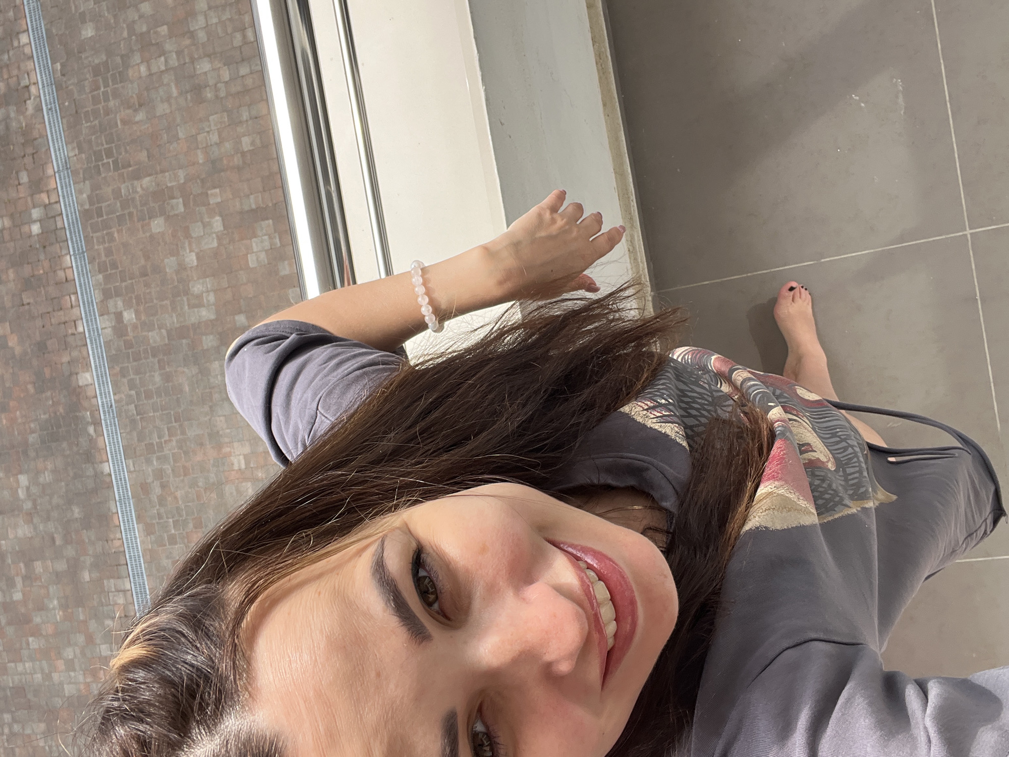 An overhead, eye-level shot captures a young woman standing on a balcony. She is wearing a gray hooded sweatshirt with a graphic design on the front and has her hair pulled up in two buns. Her right arm is extended, showing a bracelet on her wrist. Her left foot is visible, and her toenails are painted black. The background shows a tiled floor and a reddish-brown brick walkway with a metal grate running across it. A metal railing runs horizontally across the frame. The overall lighting suggests it is daytime.