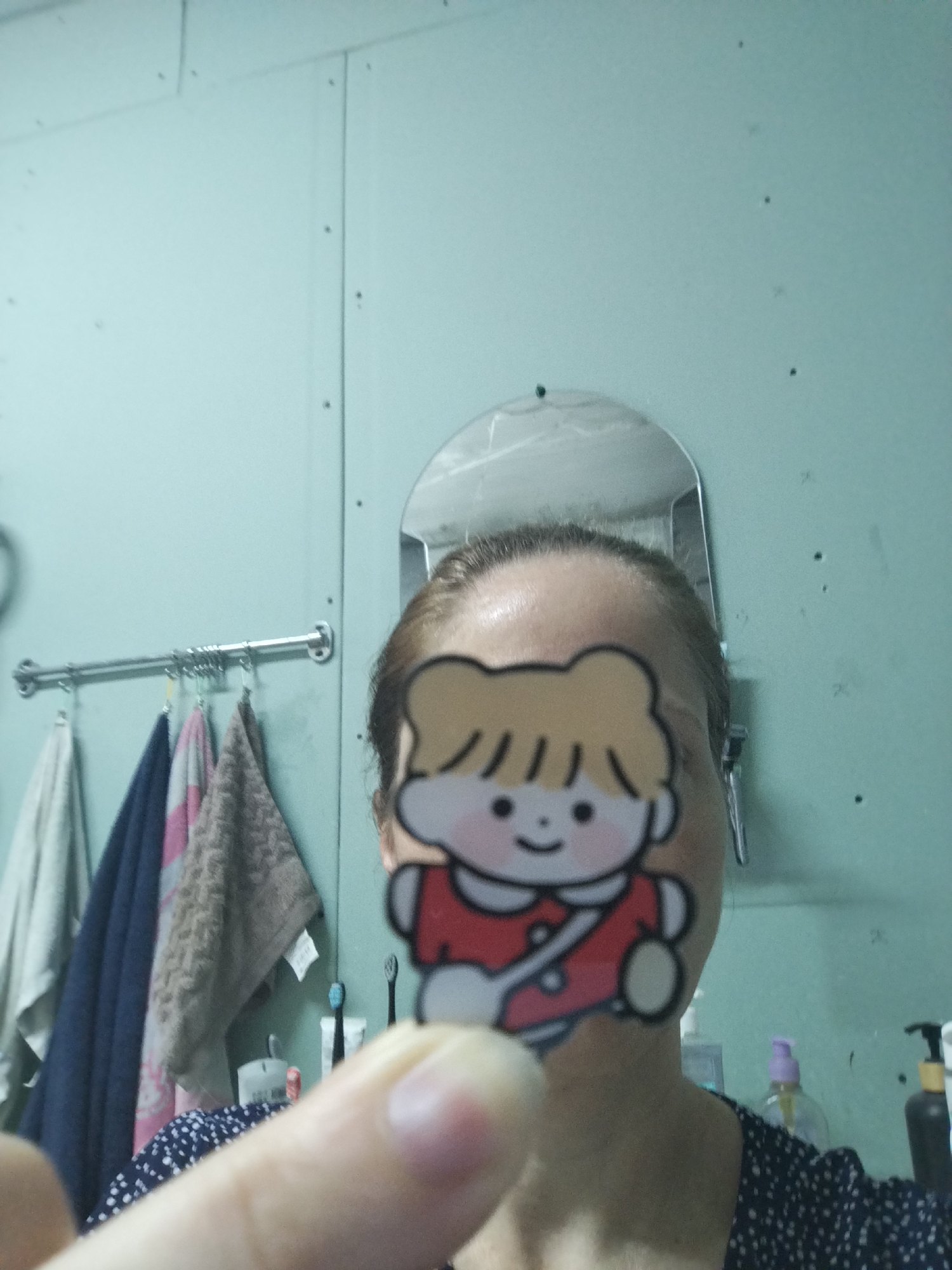 A selfie is taken in a bathroom setting. The photographer's face is partially obscured by a sticker of a cartoon child. The background includes a light blue wall, a mirror, a towel rack with folded towels, and a shelf with toiletries, including toothbrushes and bottles of liquid. The photographer is wearing a dark, polka-dotted shirt. There is no identifiable information indicating the location as Zavodoukovsk, Russia.