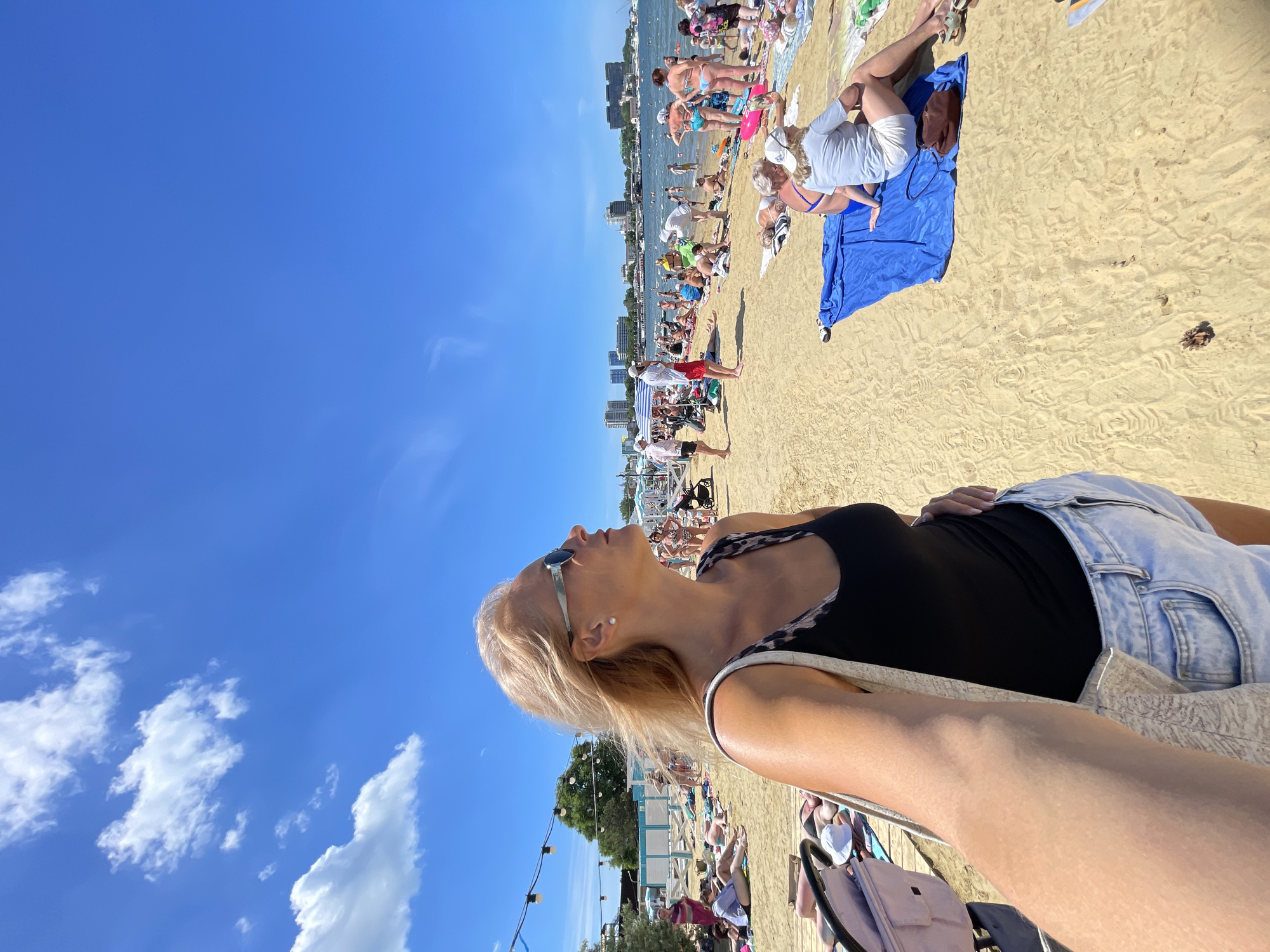 A woman with blonde hair, wearing sunglasses and a black tank top with a leopard print trim, stands on a sandy beach. She is taking a selfie, with her arm and shoulder visible in the foreground. The beach is populated with many people sunbathing and enjoying the water. In the background, there are buildings and the sea under a bright blue sky with scattered clouds. The scene appears to be a crowded public beach on a sunny day.