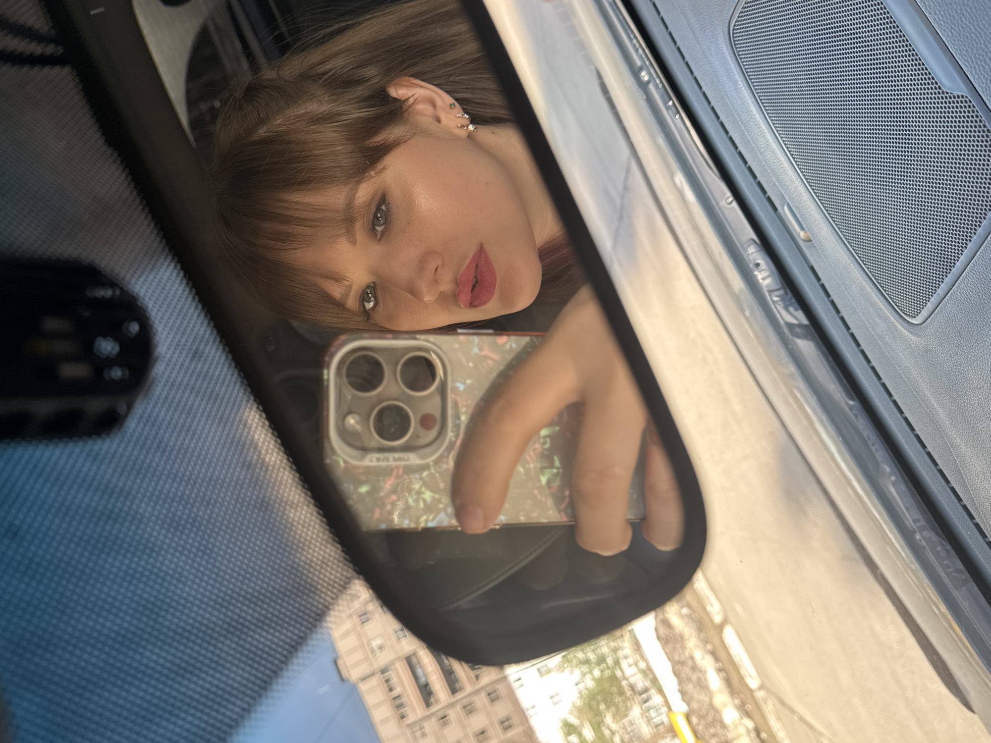A young woman is captured in a selfie reflected in a car's rearview mirror. She has light brown hair with bangs, light makeup including eyeliner and pink lipstick, and multiple small earrings in her left ear. Her expression is neutral with a slight pout, and she looks directly at the camera.
She holds a smartphone, likely an iPhone due to its three prominent rear camera lenses, which is encased in a clear, iridescent, or patterned cover. Her hand and fingers are visible gripping the phone. The reflection also shows parts of the car's interior, including a textured speaker grill on the dashboard and the car's windshield.
Outside the vehicle, reflections of urban buildings with multiple windows are visible in the mirror and through the windshield, suggesting a city environment during daytime on a clear day. The activity depicts the woman taking a self-portrait while seated inside a vehicle. No discernible text is visible. The scene is set in Chaikovsky, Russia.