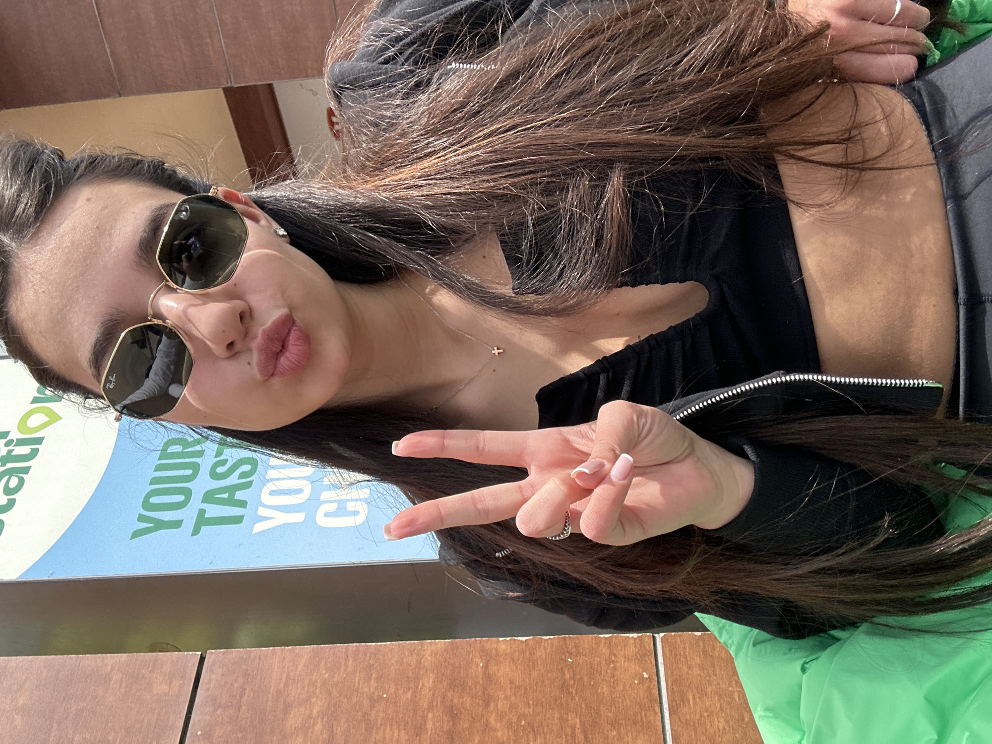 A woman is posing for a photo, wearing sunglasses and making a peace sign. She is wearing a black top and a necklace with a cross pendant. She is sitting near a sign that reads "YOUR TASTE YOU CH." The location appears to be a sunny outdoor area. The photo may have been taken in Plovdiv, Bulgaria, but this cannot be confirmed from visual information alone.