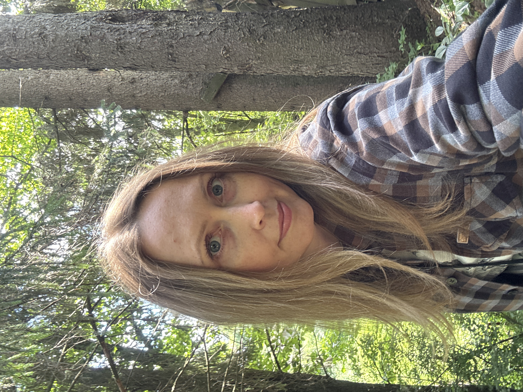 In the city of Chaikovsky, Russia, a woman with long blonde hair and blue eyes is taking a selfie outdoors. She is wearing a plaid shirt and is surrounded by trees. The sun is shining through the leaves, creating a dappled light effect. The image is a close-up of the woman's face and upper body, with the natural environment filling the background.