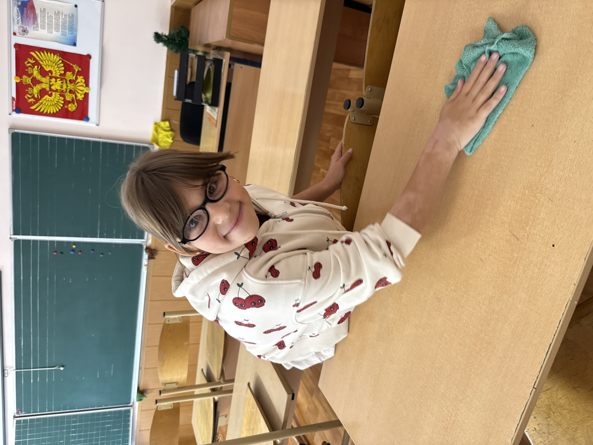 A young girl wearing glasses and a white hoodie with a cherry print is cleaning a desk in a classroom with a green microfiber cloth. The classroom appears to be in Rusanovo, Russia, with a blackboard and Russian flag visible in the background. The girl is smiling and focused on her task. The desks are made of wood and arranged in rows. The overall environment suggests a typical school setting.