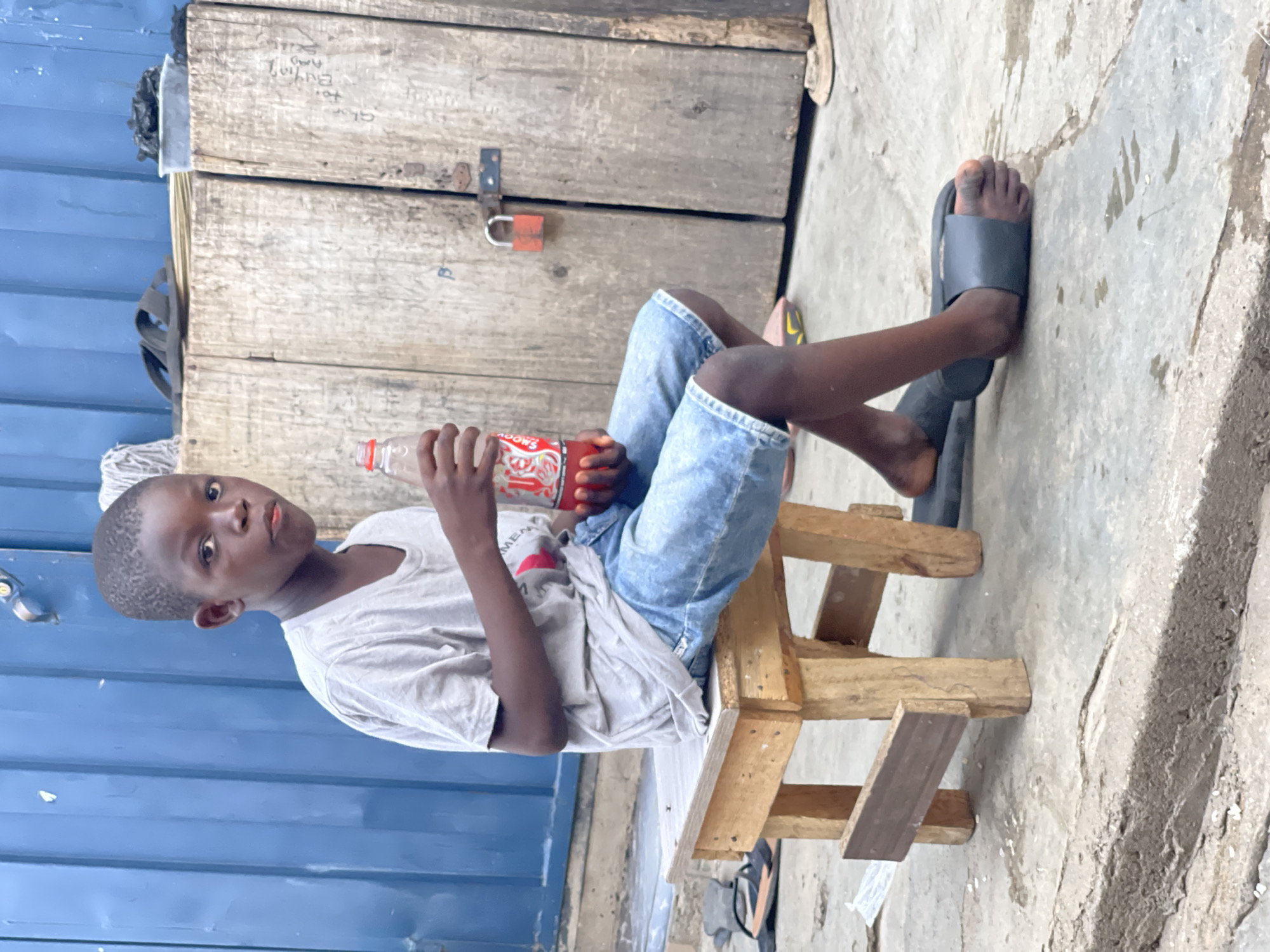 A young boy is sitting on a small, wooden stool with a Coca-Cola bottle in his hands. He is wearing a light gray t-shirt and denim shorts. His feet are adorned with black sandals. The boy is positioned in front of a corrugated blue metal wall to the left and a wooden structure that appears to be a storage unit or shed with a locked padlock. The ground is concrete. The location is Nyanya, Nigeria.