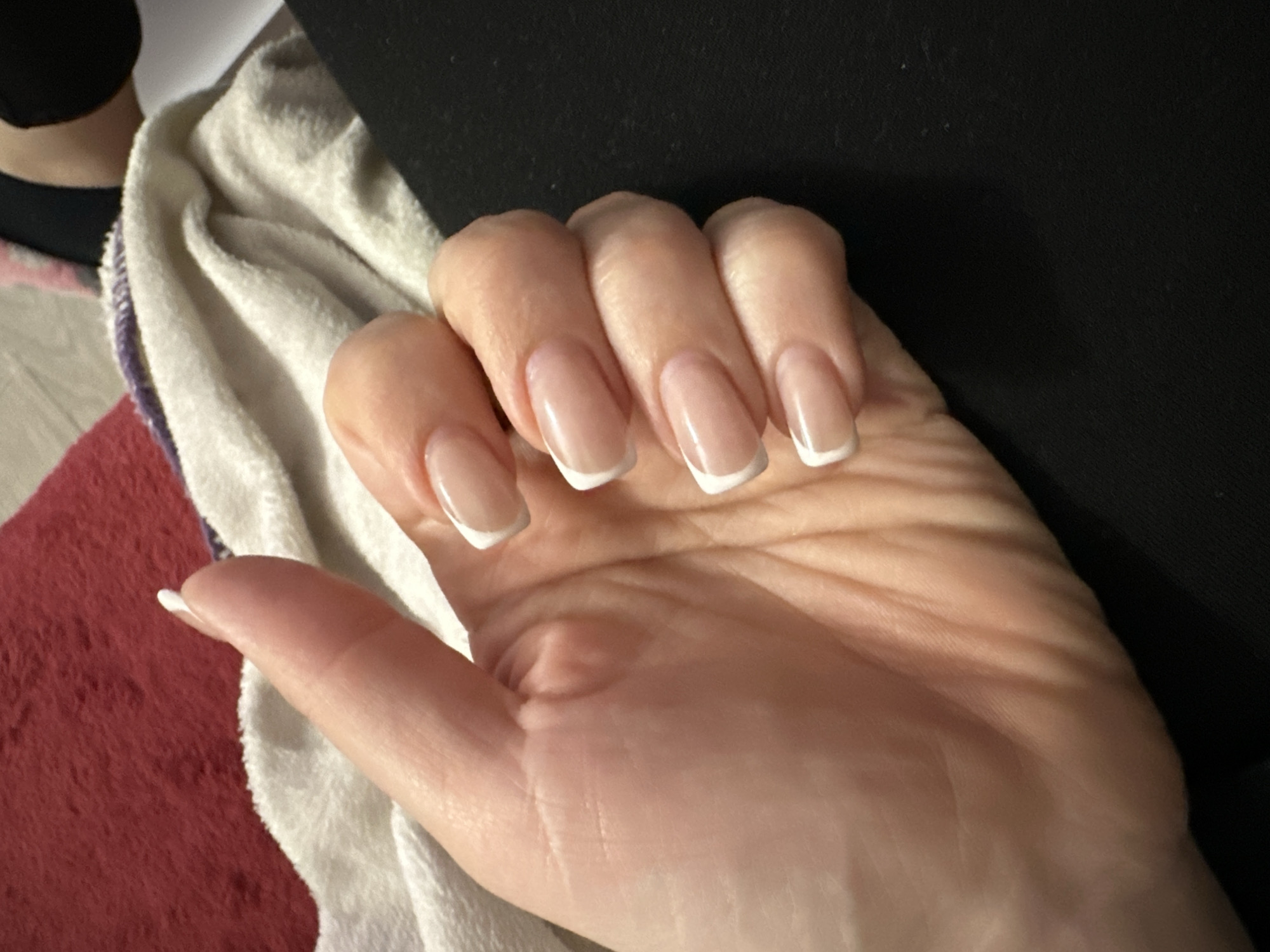 A close-up photograph shows a right hand with a fresh, clean French manicure. The nails are long and well-shaped, with a natural pink base and a crisp white tip. The hand is resting on a surface with textures of soft white terry cloth and a smooth dark material, possibly black fabric or a dark-colored surface. In the background, a glimpse of a red fuzzy rug is visible. There is no evidence to suggest the location is Sofia, Bulgaria.