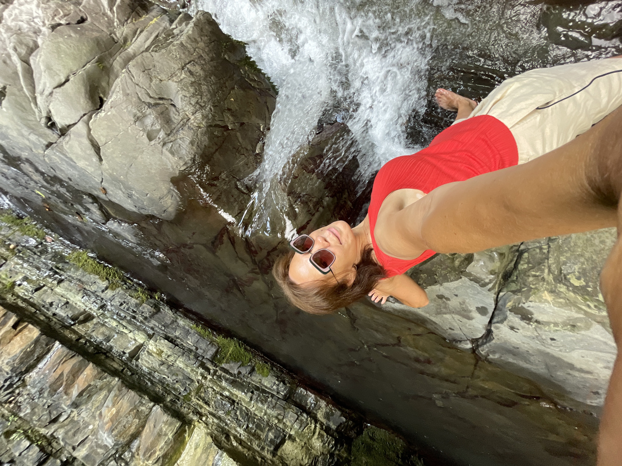 A woman with sunglasses and a red top is relaxing in a natural pool formed by a waterfall. She is lying on her back with her head tilted up, looking towards the camera. Her legs are extended, and one foot is visible submerged in the clear water. The surrounding rocks are wet and appear to be layered, with some patches of green moss growing on them. The waterfall is cascading down the rocks, creating a foamy white water effect. The overall scene suggests a peaceful and refreshing moment in nature, likely during a warm day. The image appears to be a selfie, taken from a low angle. No text is visible in the image. The location context provided is Bolshoy Sochi, Russia.