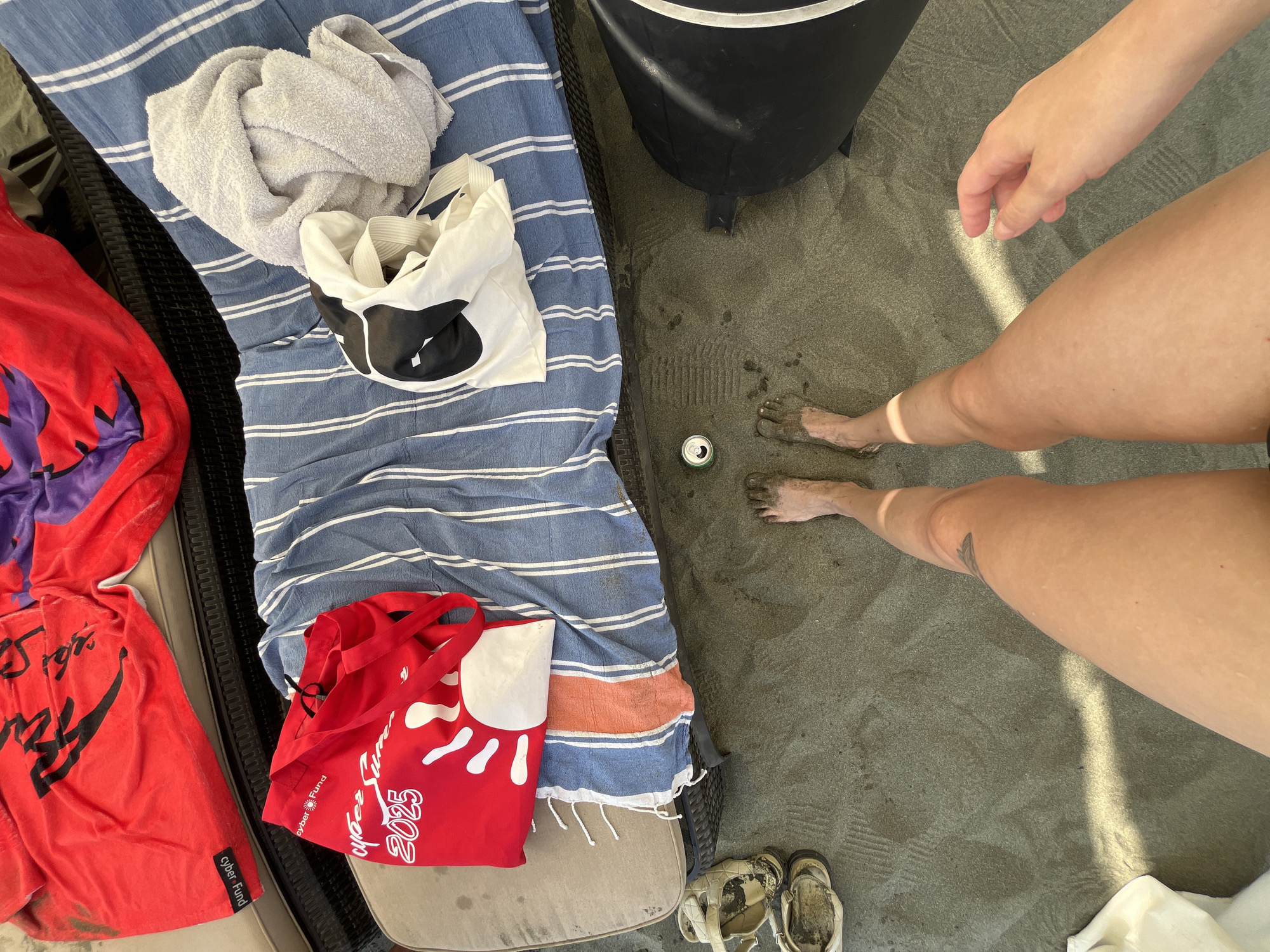 A first-person perspective shot captures a beach scene in Agios Tychona Community, Cyprus. The image is dominated by sand, with a beach lounge chair in the background covered in striped blue and white beach towels, a red beach bag, and a grey towel. A pair of dirty sandals are placed on the sand to the left, and a black container is visible on the right. In the center, a person's bare legs and feet, caked with sand, are visible. Their left leg shows a tattoo on the thigh. A hand extends from the right side of the frame, partially obscuring the lower right corner. The overall impression is one of a relaxed day at the beach.