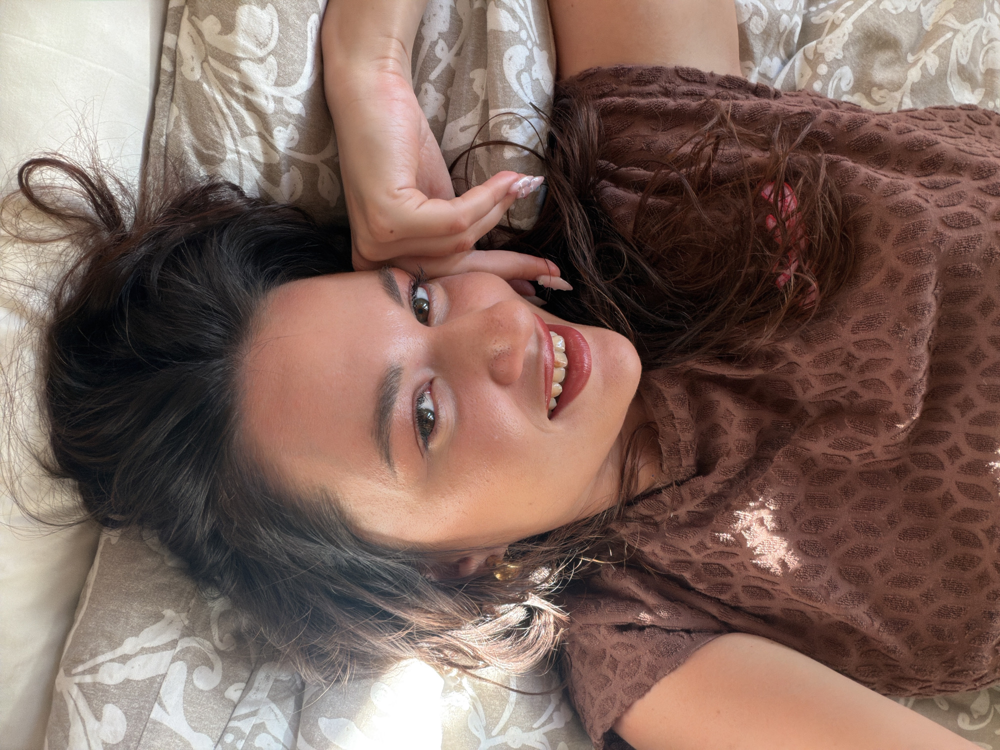 An overhead shot shows a young woman lying on her back on a bed. Her dark brown hair is spread out around her head. She is wearing a brown textured t-shirt and has her right hand resting on her cheek. Her mouth is open, revealing her teeth and red lipstick. Her eyes are looking at the camera. The bed is covered with a patterned duvet. The lighting suggests it is daytime, with sunlight casting shadows on her face and t-shirt. There is no indication of the location being in North Kuta, Indonesia.