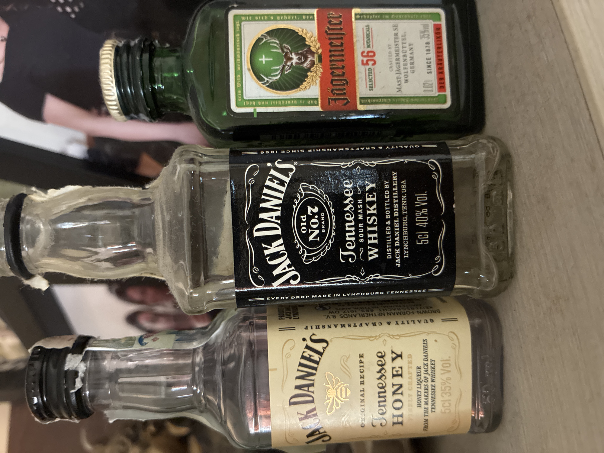 The image displays three miniature, likely empty, liquor bottles, arranged horizontally and stacked one above the other in an indoor setting. A blurry reflection or image of a person, possibly a female with dark hair and a black top and a white wristband, is visible in the background.
The top bottle is a green glass Jägermeister bottle with a gold/silver cap. Its label features the Jägermeister brand name, a stag's head with a cross between its antlers, and text identifying it as a "Kräuterlikör" (herbal liqueur), crafted by "MAST-JAGERMEISTER SE WOLFENBÜTTEL, GERMANY," with a volume of 2cl and 35% alcohol by volume. White, fuzzy residue or dust is visible around the cap.
The middle bottle is a clear glass Jack Daniel's Old No. 7 Tennessee Whiskey bottle, featuring a black label and black cap. The label prominently displays "JACK DANIEL'S Old No.7 BRAND Tennessee SOUR MASH WHISKEY," along with "DISTILLED & BOTTLED BY JACK DANIEL DISTILLERY LYNCHBURG, TENN. USA." It specifies a volume of 5cl and 40% alcohol by volume, and notes "QUALITY & CRAFTSMANSHIP SINCE 1866" and "EVERY DROP MADE IN LYNCHBURG TENNESSEE."
The bottom bottle is a clear glass Jack Daniel's Tennessee Honey bottle, also with a black cap. Its tan/gold label features a bee graphic and text that reads "JACK DANIEL'S ORIGINAL RECIPE Tennessee HONEY HONEY LIQUEUR FROM THE MAKERS OF JACK DANIEL'S TENNESSEE WHISKEY," with a volume of 5cl and 35% alcohol by volume. It also states "QUALITY & CRAFTSMANSHIP" and that it's "PRODUCED BY BROWN-FORMAN NETHERLANDS B.V."
All bottles appear dusty, suggesting they have been stored for some time. No specific activity is taking place; the bottles are simply displayed. The lighting is ambient indoor light, and no specific time of day is indicated. The scene is located in Varna, Bulgaria.