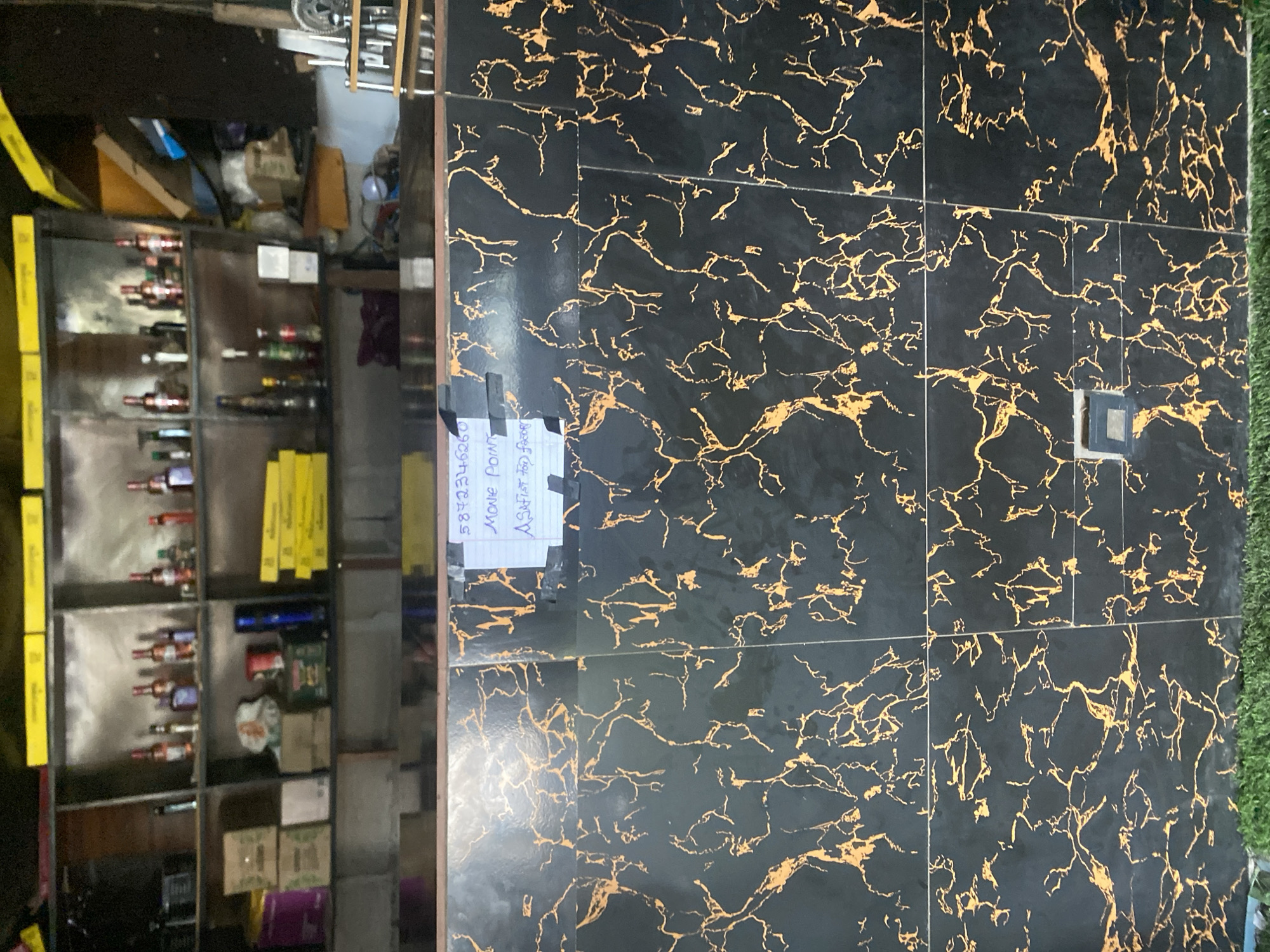 This media file depicts a retail setting, possibly a liquor store or convenience store, in Jalingo, Nigeria. The foreground is dominated by a black marble-patterned wall with gold veining. Affixed to this wall are two pieces of white paper with handwritten text. The left paper displays the numbers "587234" and "346260" vertically. The right paper contains the words "MOVE POINT" and "ASHTAR FOR".
To the left of the marble wall is a shelving unit filled with various items. The shelves contain rows of bottles, many appearing to be liquor bottles, with dark liquid. Some shelves also hold boxes and other merchandise. The overall lighting suggests an indoor environment, likely during daylight hours given the context of a retail space. The arrangement of goods indicates a functional retail space, possibly with a focus on beverages. No people are visible in the frame. The scene appears to be a typical commercial environment.