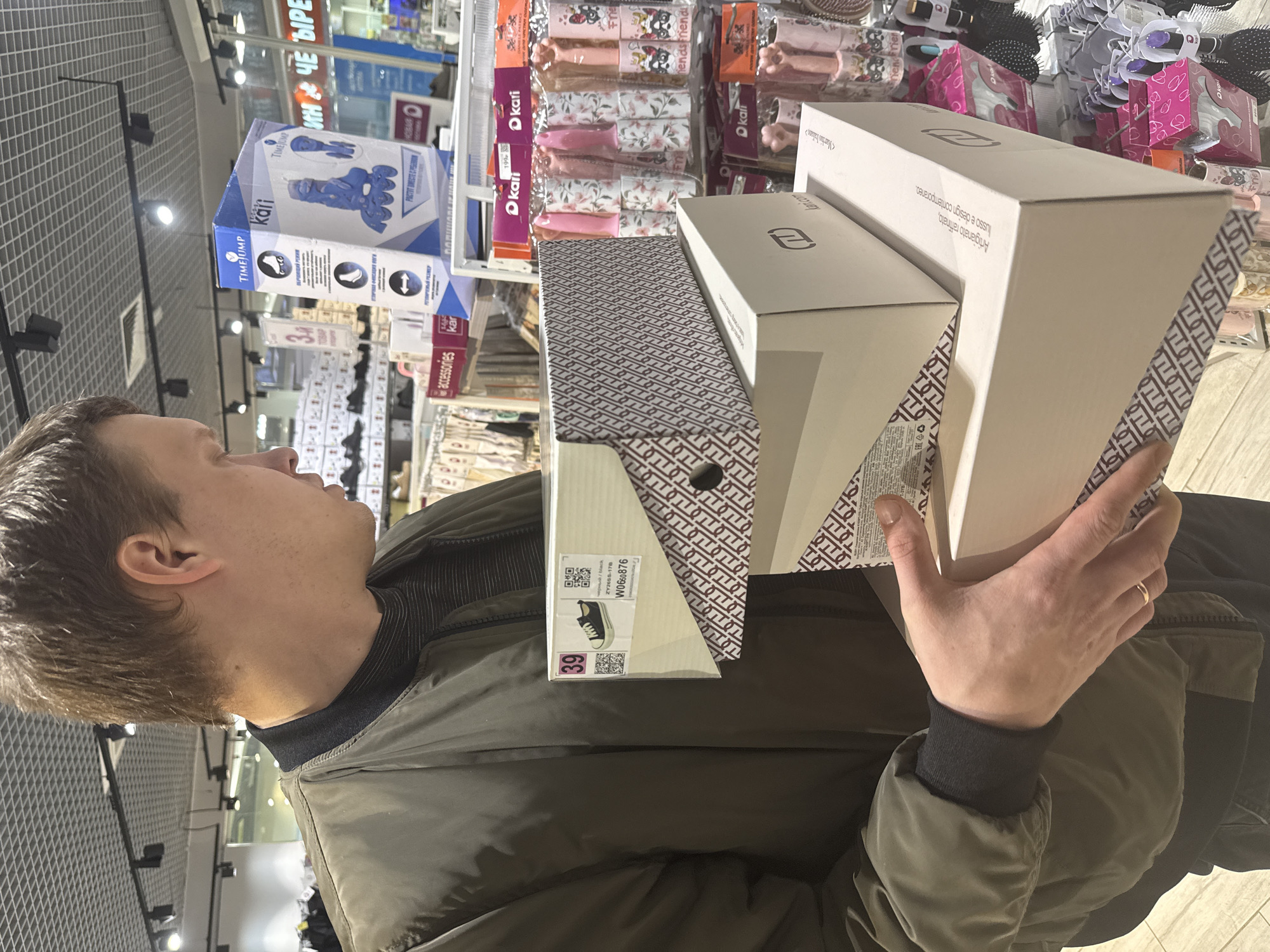 A person is holding three shoe boxes in a store. The boxes are white with black text and patterns. The person is wearing a green jacket and has short brown hair. The boxes are stacked on top of each other. The topmost box is the tallest. Shelves with various items, including shoes and accessories, are visible in the background. Signs and labels in Russian and English can be seen on the shelves. The location is identified by labels and signs containing "kari" and "TIMEJUMP". OCR indicates the location is in the city of Dmitrov, Russia. The shoe size is marked as 39.