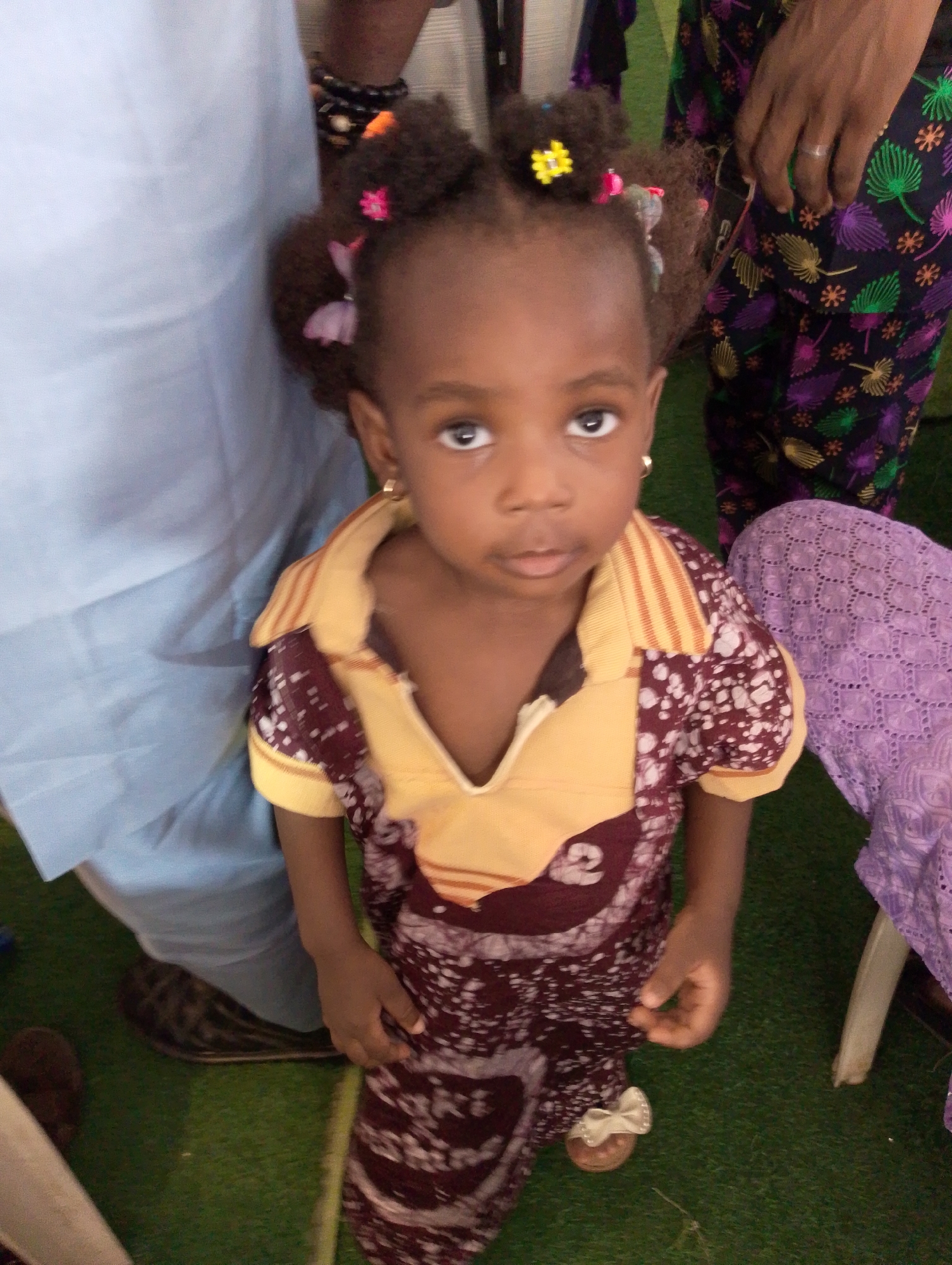 A young child with dark skin is the primary subject of this image. She is wearing a dress that has a pattern of maroon and white, with a yellow collar and short sleeves. Her hair is styled in small puffs adorned with colorful flower clips. The child is wearing small gold earrings.
Two adults partially appear in the image, standing on either side of the child. The person on the left is wearing a light blue garment. The person on the right is wearing pants with a vibrant floral pattern.
The setting appears to be indoors, with a green carpet visible beneath the subjects' feet. A white plastic chair is partially visible in the lower right. The image was taken in Dushepe, Nigeria.