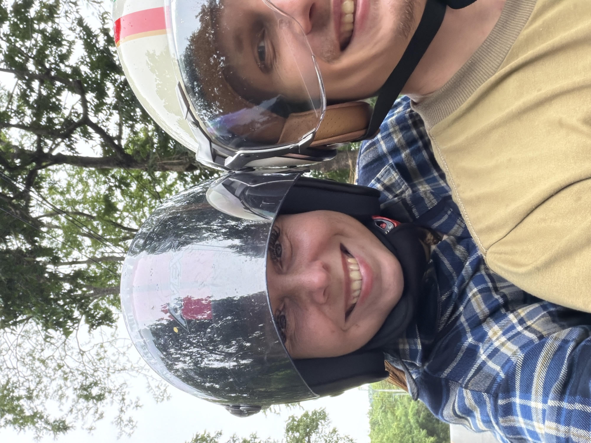 Two individuals are wearing helmets and riding on a motorbike. The person in the foreground, on the left, is a woman with brown hair, wearing a blue and white plaid shirt and a black helmet with a reflective visor. She is smiling broadly. The person behind her, on the right, is a man with short hair, wearing a tan shirt and a cream-colored helmet with a clear visor. He is also smiling. They appear to be on a road with trees and foliage in the background, suggesting a rural or natural setting. The weather appears overcast. The image was taken in Samoeng Tai, Thailand.
