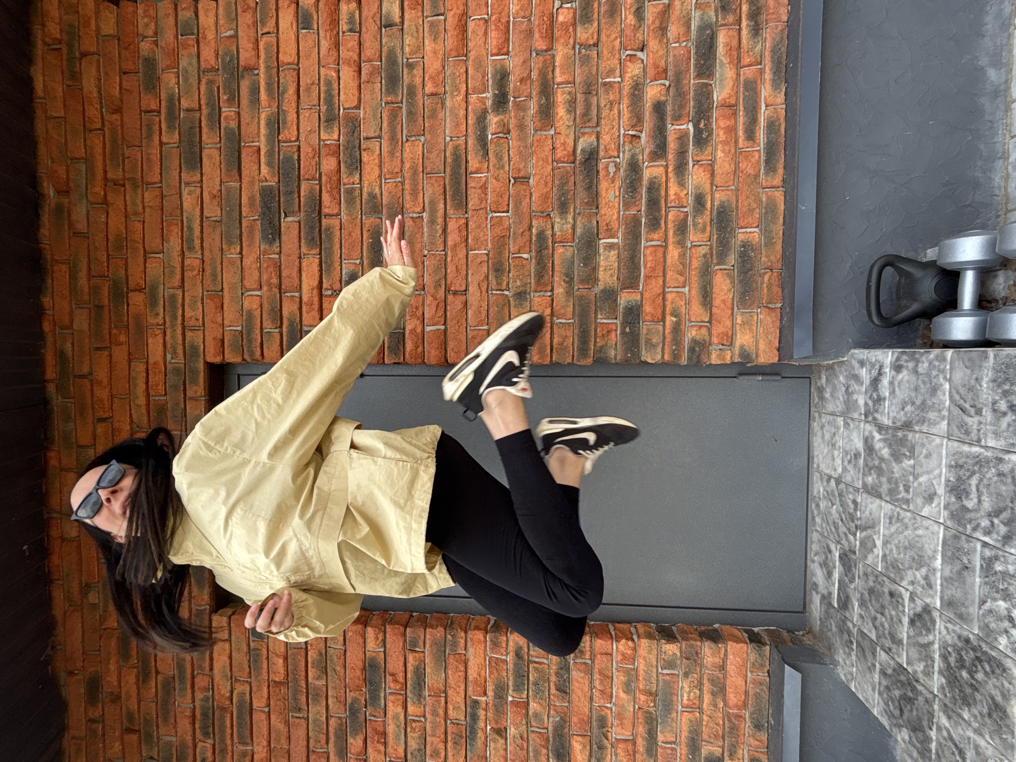 The image captures a person in a dynamic, mid-air pose against a textured brick wall, rotated 90 degrees clockwise from its likely original orientation.
The primary subject is a woman with long, dark hair, wearing dark sunglasses that obscure her eyes. She is dressed in a light yellow or beige long-sleeved jacket and black leggings. Her footwear consists of black and white athletic shoes, with the iconic Nike swoosh visible on them. She appears to be in motion, possibly jumping or floating, with her right arm extended upwards and her legs bent at the knees, elevated off the ground.
The background consists of two distinct wall sections. To the left, there is a prominent vertical wall constructed from reddish-brown bricks with dark, burnt-like streaks, suggesting an industrial or rustic aesthetic. To the right of the brick wall, a solid dark gray wall or panel extends upwards. Below this gray wall, and extending further to the right, is a lighter gray, textured floor or platform.
On this gray platform, positioned in the bottom right corner of the image, several fitness items are visible: a black kettlebell and a stack of silver dumbbells, indicating a gym or exercise-related setting. The lighting is bright, suggesting the photo was taken during the daytime. No specific emotions are discernible due to the sunglasses and dynamic pose. The location context provided is Chaikovsky, Russia.