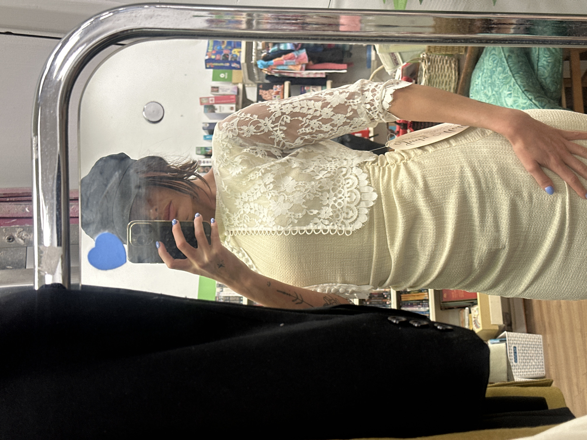 The image shows a reflection in a mirror, presumably of a person taking a selfie. The person is wearing a light-colored dress with lace sleeves and is holding a smartphone in their hand. The background of the reflection shows a cluttered space with various objects, including books, storage boxes, and other items. In the foreground is a black garment, possibly a coat or jacket. No specific details in the image allow for the location to be identified as Tunbridge Wells, United Kingdom.