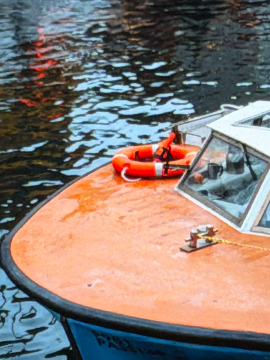 The image shows the bow of an orange boat in the water. A life preserver with red and white stripes is visible on the deck of the boat. A mooring cleat is also present. The boat is in water that reflects red and white lights. The boat has blue lettering on its side. There are no people visible in the image. The scene is a close-up of the boat and its immediate surroundings. The location is the city of Kafr Yasif, Israel.