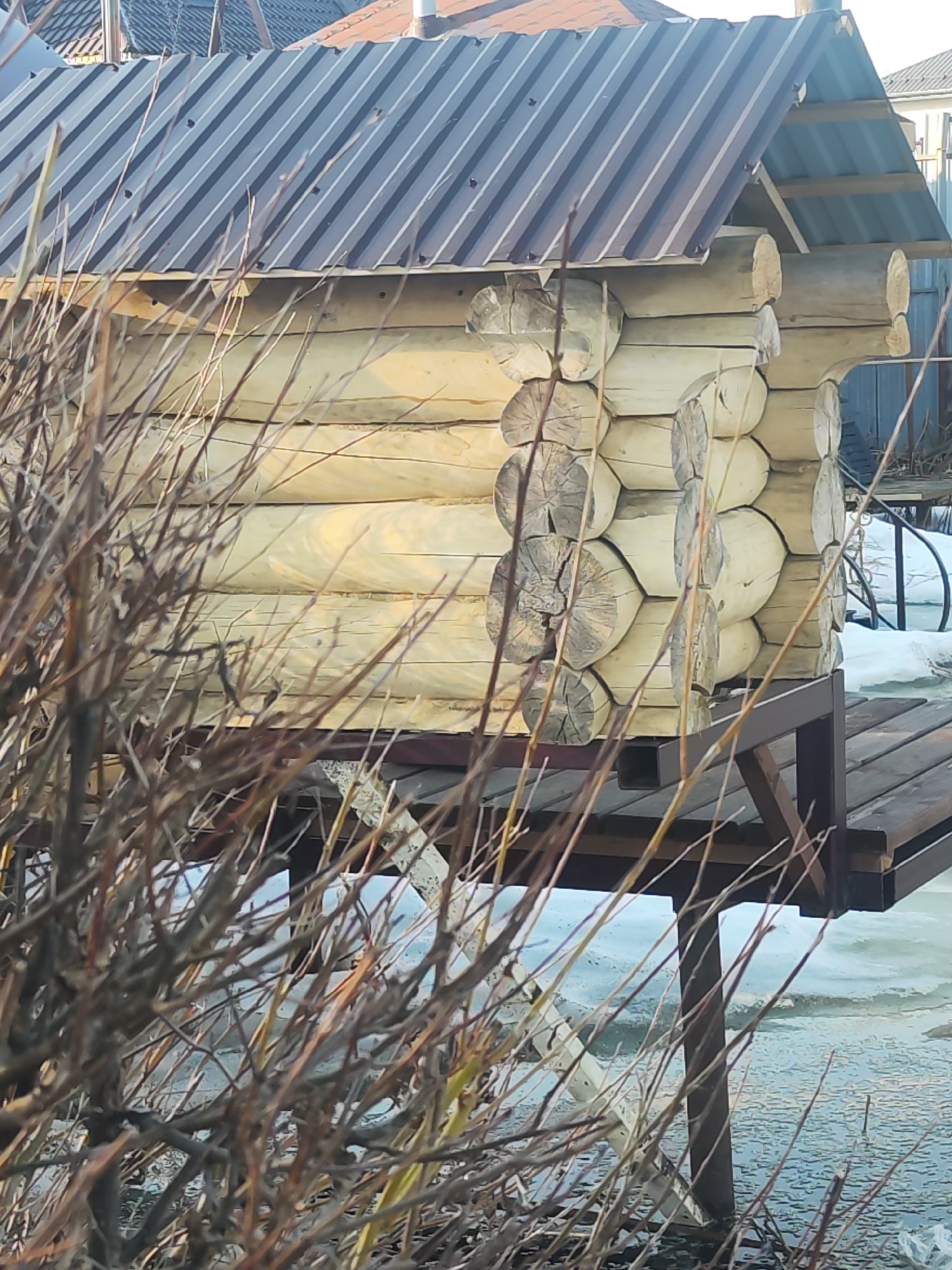 A small, unfinished log cabin with a corrugated metal roof sits on a wooden platform supported by metal legs, located in Chaikovsky, Russia. Bare branches obscure much of the lower portion of the cabin. Patches of snow and ice are visible around the base of the structure. In the background, a partially visible structure with a red roof is present.