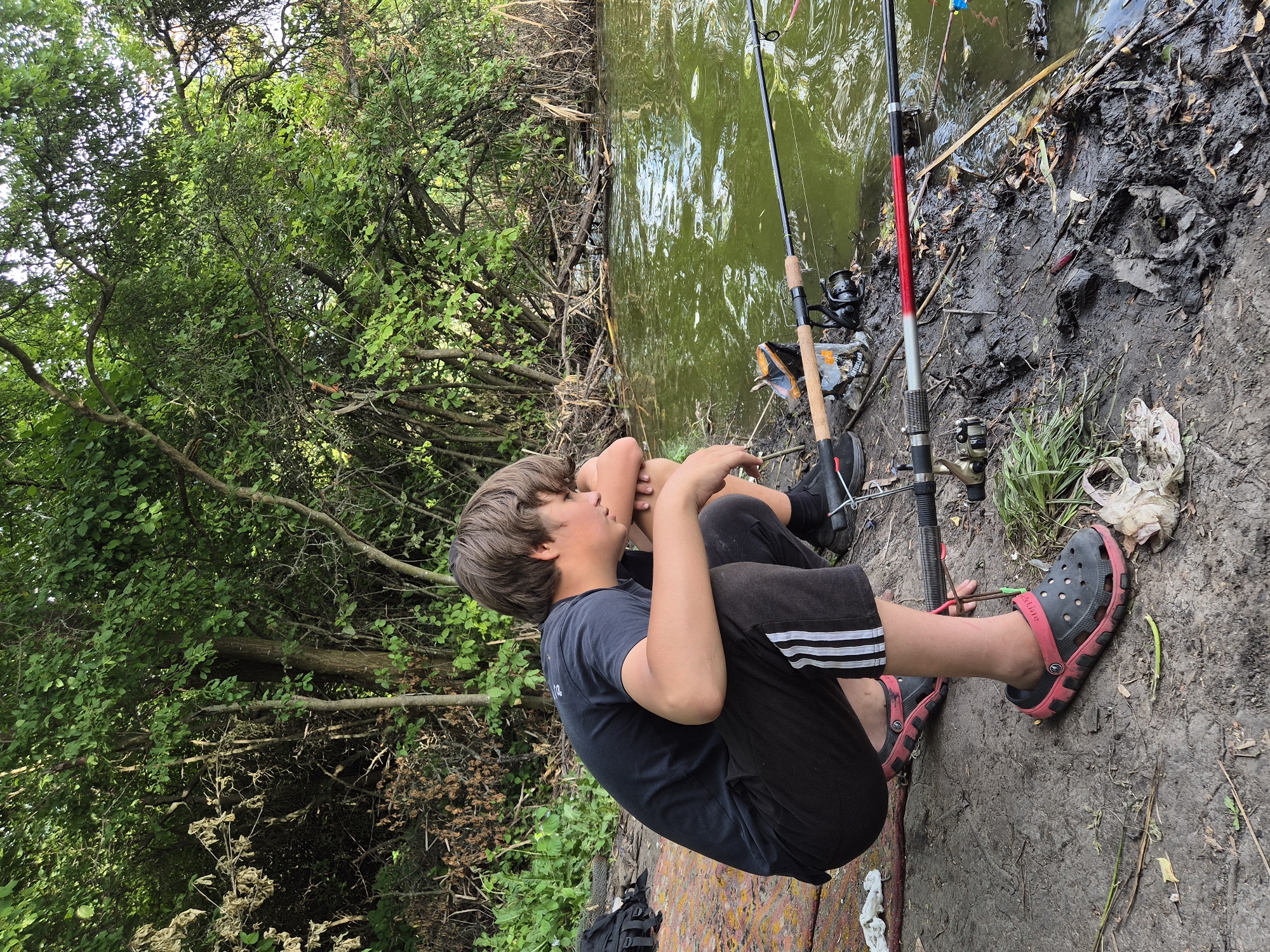 A young boy with brown hair sits on the muddy bank of a murky green body of water, appearing bored or tired. He is wearing a black t-shirt with a graphic on the left chest, black shorts with white stripes down the side, and black and pink crocs. His arms are crossed over his chest and his head is resting on his arms. Two fishing rods are propped up on the bank. One rod is black with a silver reel and is partially obscured. The other rod is black with a red and white accent on the upper portion and a silver reel. The background consists of dense green foliage from trees and bushes, and the ground is muddy with debris. The scene suggests a fishing trip on a partly cloudy day. The location is Sozonivka, Ukraine.