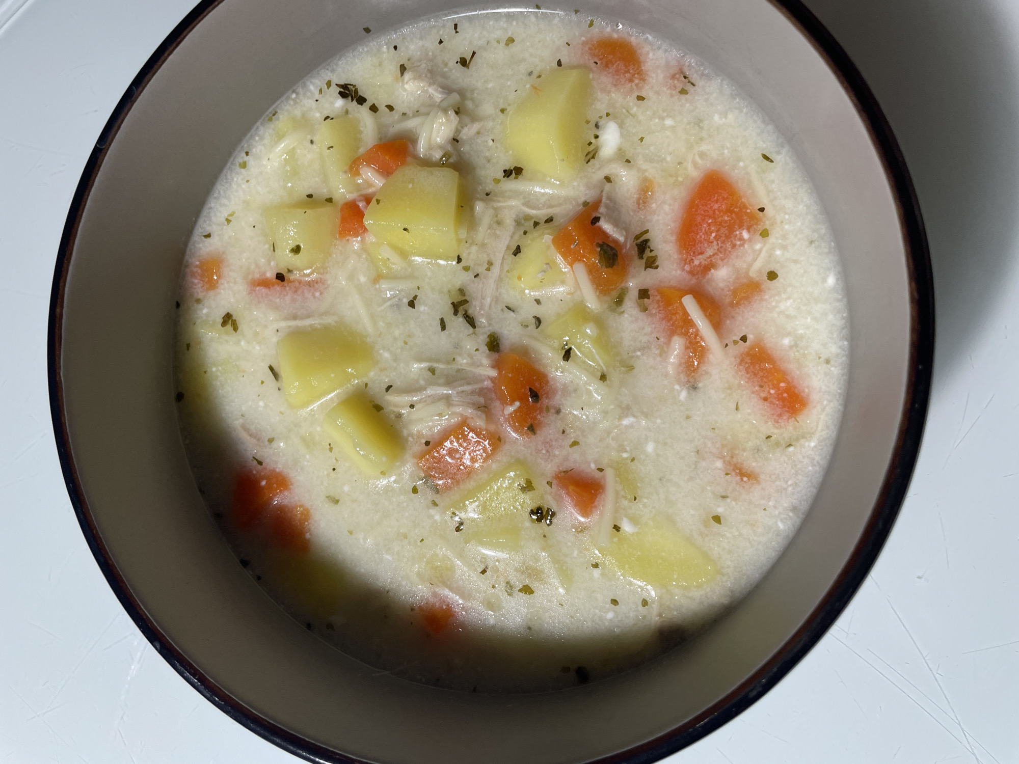 A close-up, top-down shot shows a bowl of chicken noodle soup, featuring diced potatoes, carrot slices, shredded chicken, and noodles in a creamy, white broth, garnished with dried herbs. The bowl has a light grey interior and a dark brown rim. The bowl sits on a white surface. The scene lacks any identifiable features indicating it's in Izhevsk, Russia.