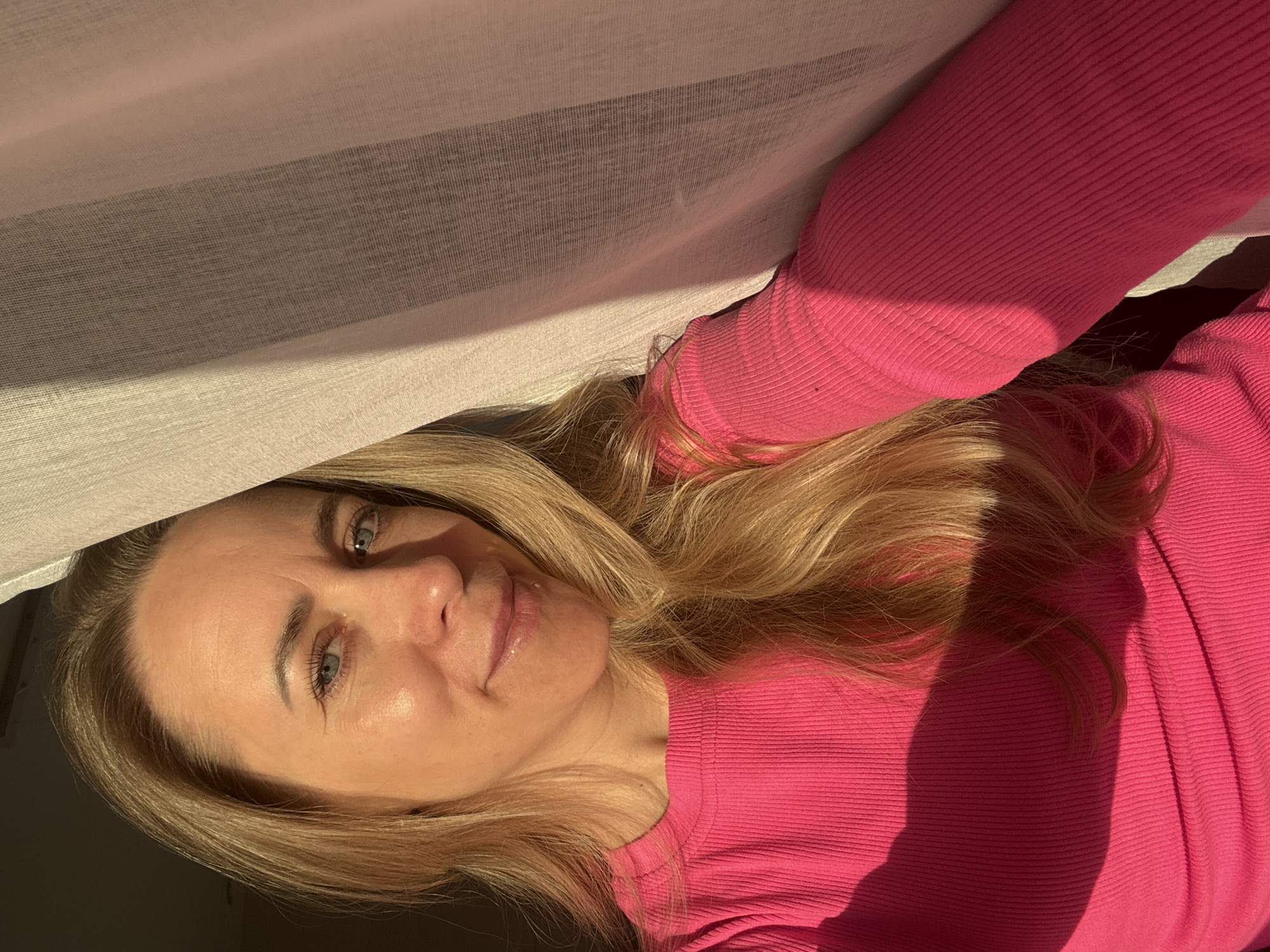 A selfie of a woman with blonde hair and blue eyes, wearing a bright pink long-sleeved shirt. She is partially obscured by a light-colored curtain. The location is unidentifiable in the city of Chaikovsky, Russia, from the given image.
