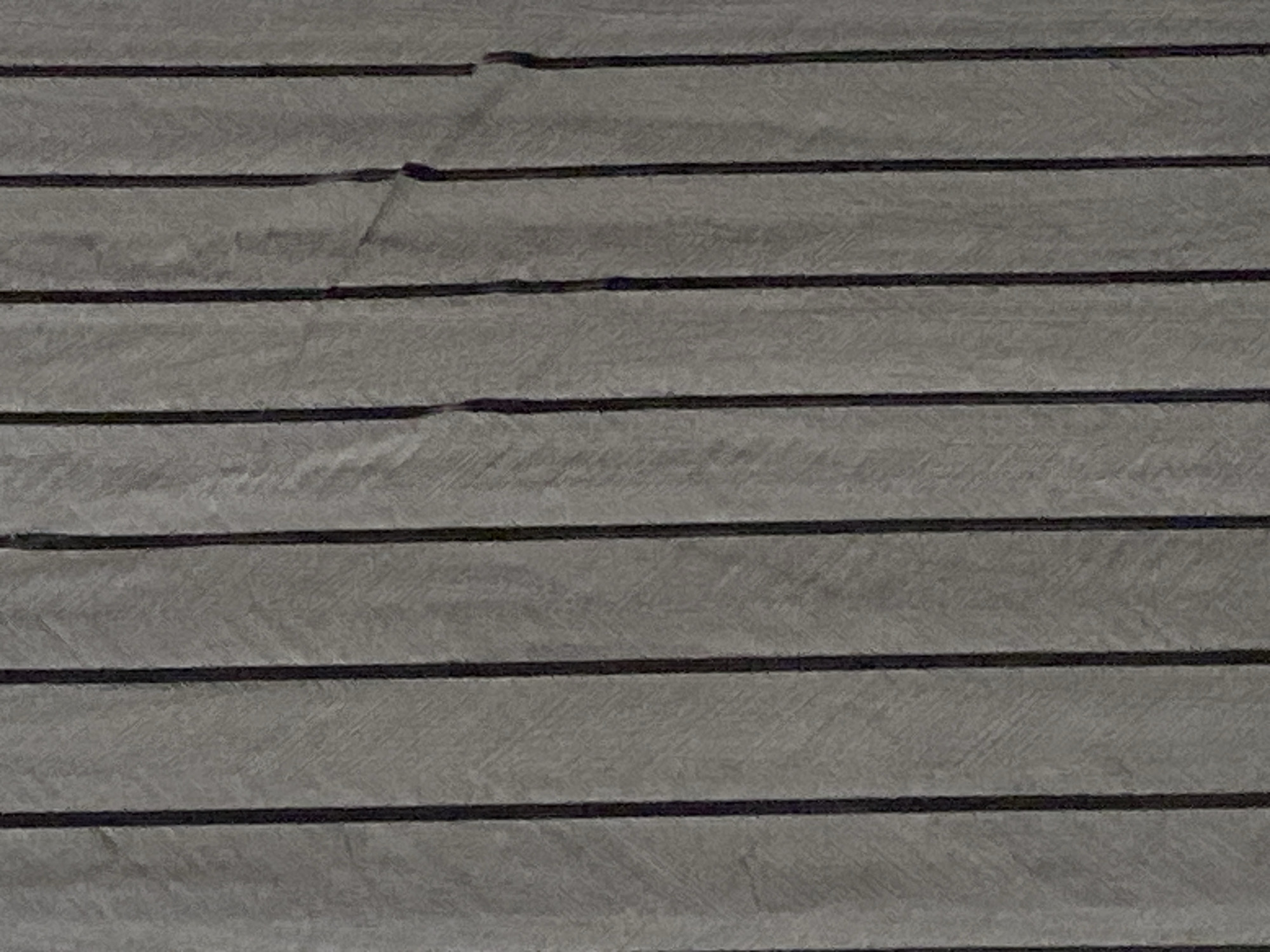 This is a close-up, full-frame photograph of a gray, textured surface with vertical black stripes. The texture of the gray material appears to be fabric or a similar woven material, with subtle diagonal lines creating a pattern within each stripe. The black stripes are of uniform width and are evenly spaced. There are minor imperfections or creases visible in the gray material between some of the stripes, particularly in the middle and right sections of the image, where the material appears to be pulled or creased. The lighting is even, with no harsh shadows, suggesting an indoor setting or overcast conditions. The image provides no information about the location of Jauro Yinu, Nigeria, as it is an abstract representation of a surface texture. There are no people, animals, or discernible objects other than the striped surface itself.