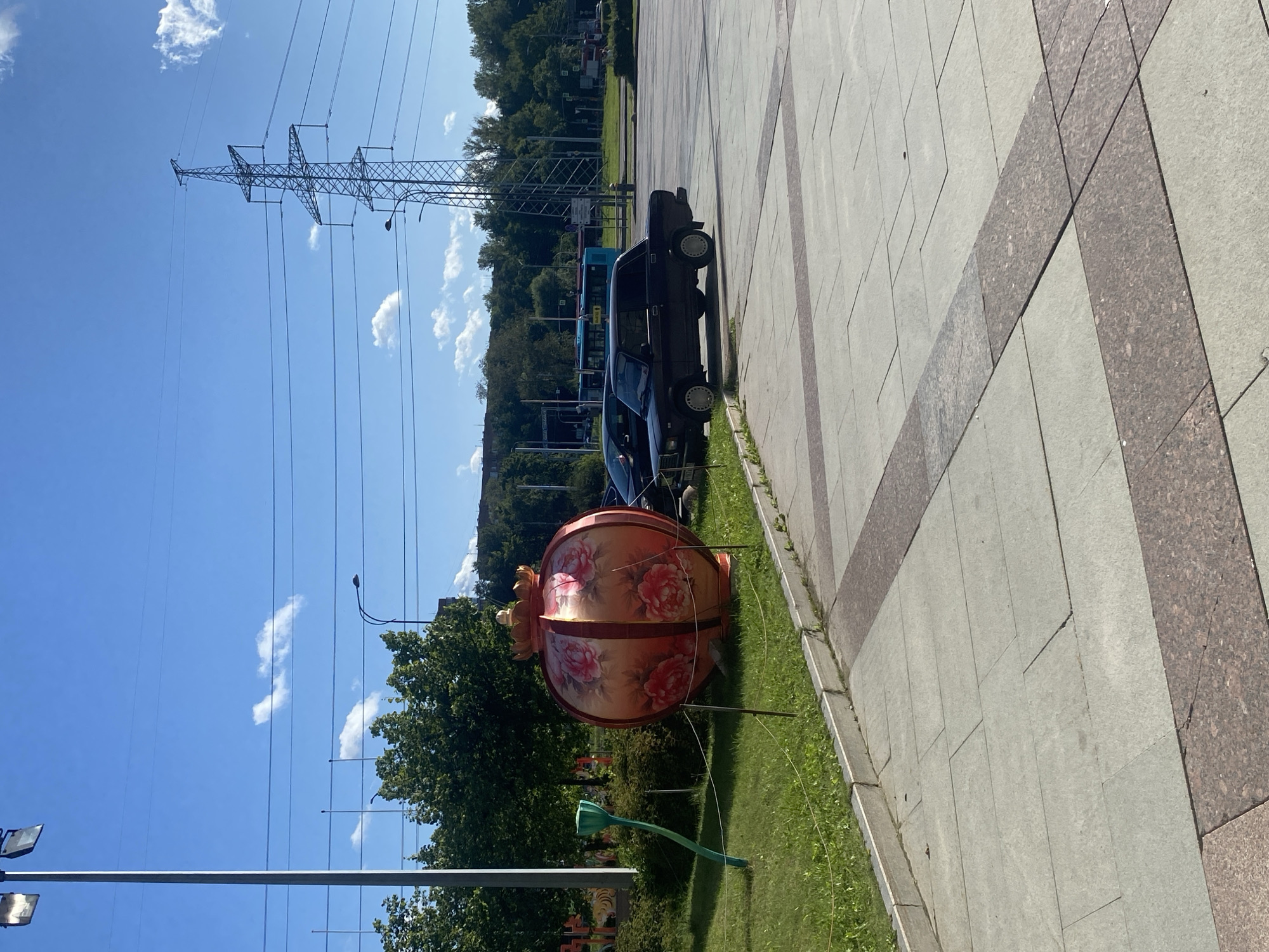 The image depicts an outdoor scene on a sunny day in St. Petersburg, Russia. In the foreground, a paved area with a pattern of light gray and reddish-brown stone tiles suggests a pedestrian walkway or plaza. To the left, a grassy area is bordered by a curb and a thin rope fence. A large, decorative, spherical lantern, painted with pink flowers, stands prominently on the grass. Adjacent to it is a green, stylized sculpture resembling a plant stem.
In the middle ground, two dark-colored cars are parked. The one on the right is a classic sedan, possibly a Lada. Behind the cars, a street with trees and a bus is visible. In the background, a large electricity pylon with multiple power lines stretches across the frame against a clear blue sky with scattered white clouds. Trees line the horizon, indicating a suburban or park-like setting within the city. The presence of the lantern and stylized sculpture suggests a public space decorated for an event or as a permanent art installation.