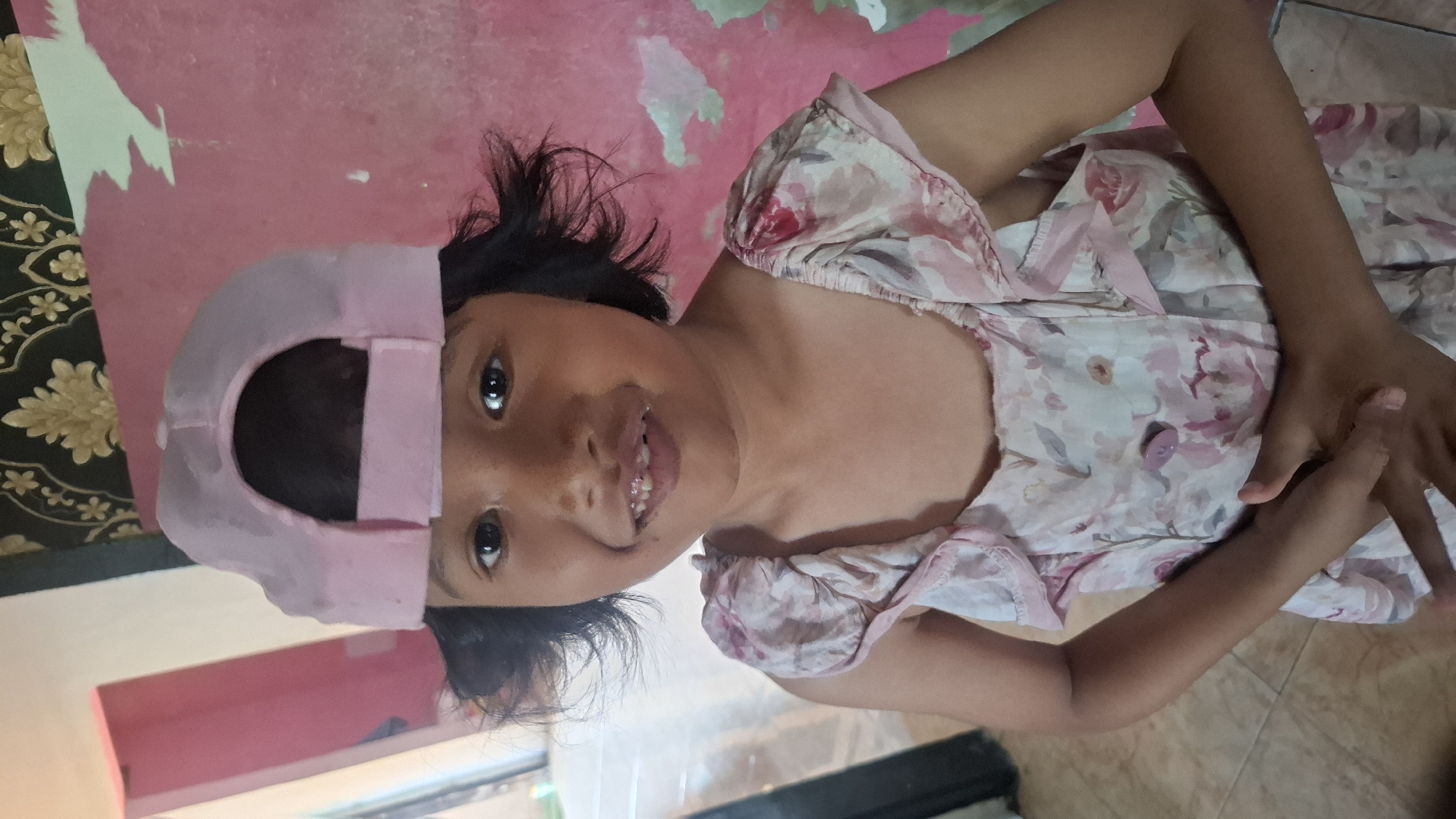 A young girl with dark hair, wearing a pink baseball cap and a floral dress, smiles at the camera. She is standing in a room with a pink wall and a patterned wallpaper in the background. The floor is tiled. Her hands are clasped in front of her. The scene is likely indoors, possibly a residential setting in Cibinong Hilir, Indonesia.