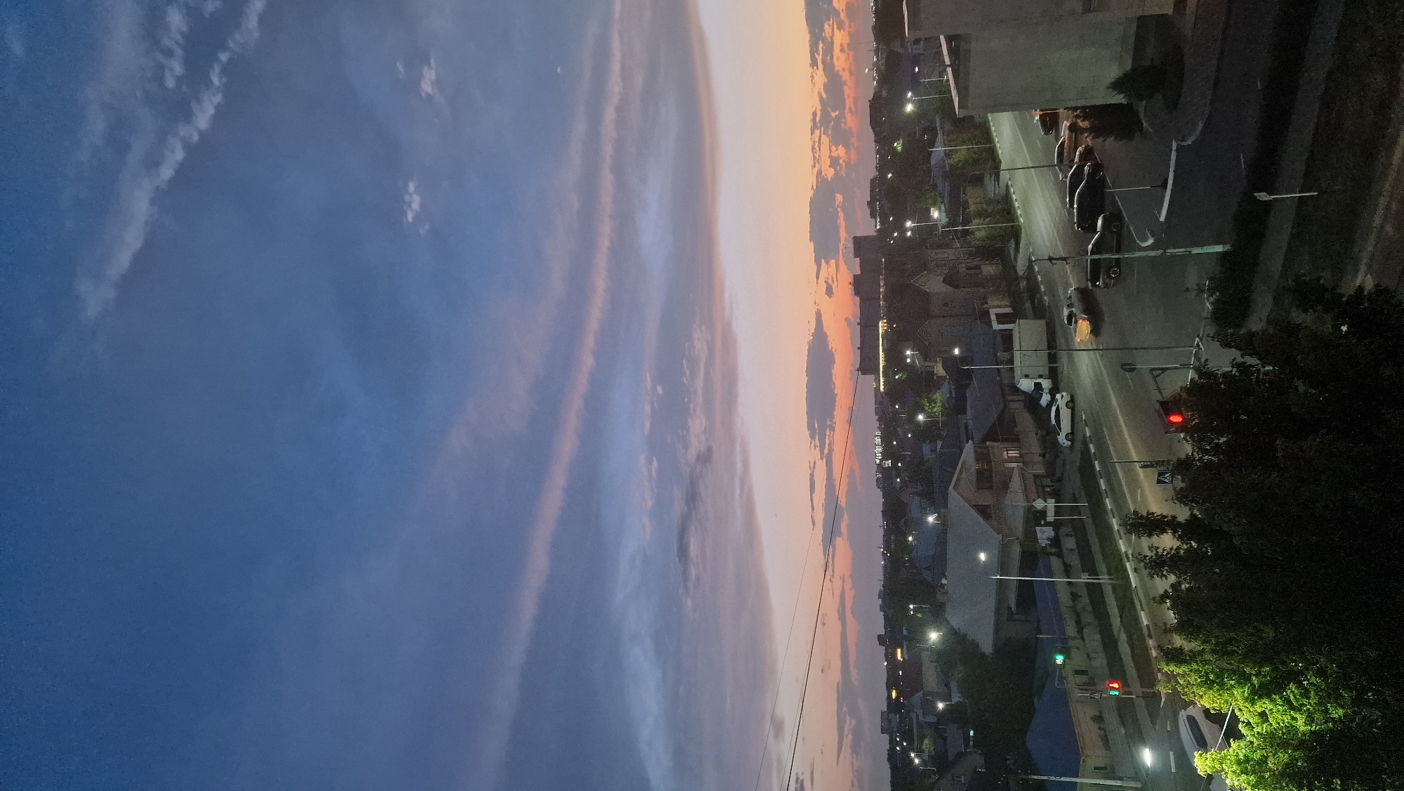 The image depicts a twilight scene of a city street in Kostanay, Kazakhstan. The sky is a gradient of deep blue at the top, transitioning through shades of purple and pink, with wispy clouds catching the last light of the setting sun. Below the sky, a cityscape is visible, characterized by houses with pitched roofs, streetlights illuminating the roads, and a few multi-story buildings in the distance.
The foreground shows a street with traffic. A white sedan and a truck are parked on the side of the road, while several cars are driving or parked on the main thoroughfare. One car's headlights are illuminated, indicating movement. Traffic lights are visible, one showing red. Dense foliage, likely trees, is prominent on the left side of the image, with some of the leaves illuminated by artificial light. The overall impression is of a residential or mixed-use urban area at dusk, with signs of everyday activity.